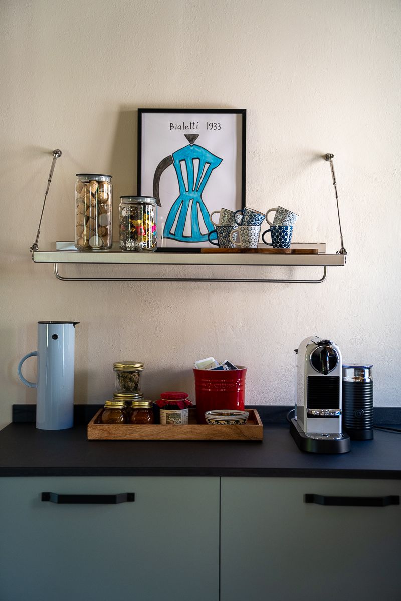 Modern kitchen shelf with Bialetti poster, glass jars, mugs, and a Nespresso machine on dark countertop.