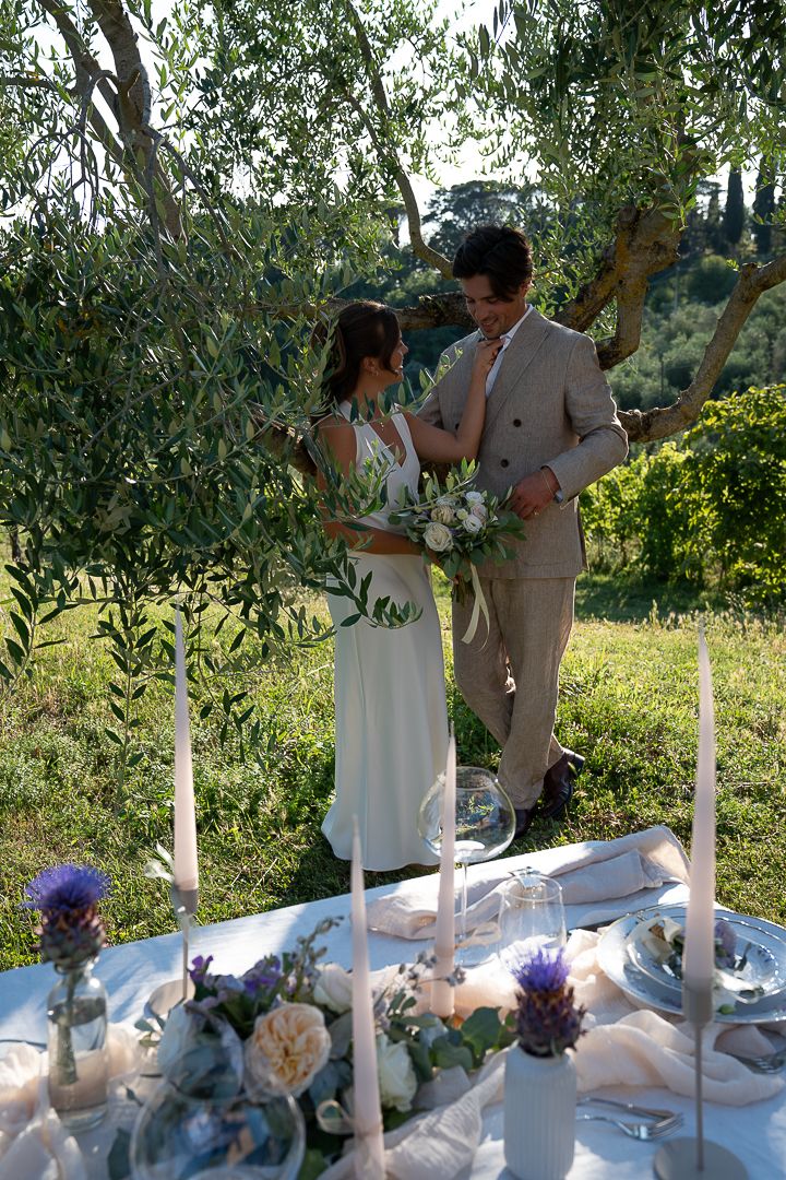 Bride and groom stand together under olive trees beside an elegantly decorated outdoor wedding table in Umbria on their elopement day.