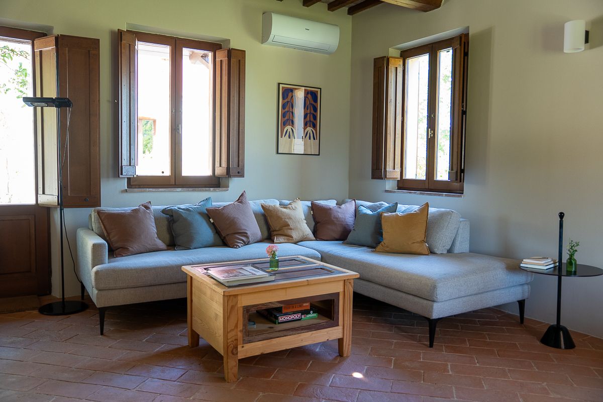 Cozy living room with a light gray L-shaped sofa, colorful cushions, wooden coffee table, and terracotta tile floors.