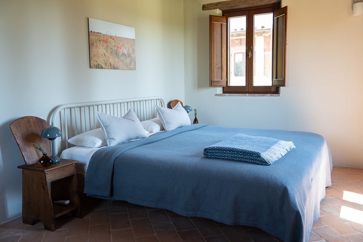 Cozy bedroom with a blue bedspread, white pillows, wooden nightstand, and rustic terracotta floor tiles near a window.