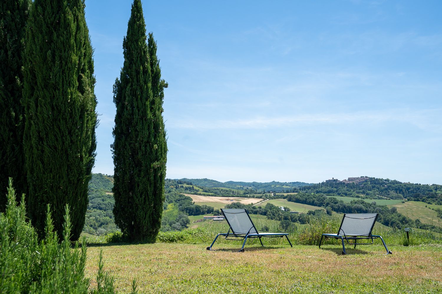 Two lounge chairs overlook rolling Tuscan hills with tall cypress trees framing a peaceful countryside vista.
