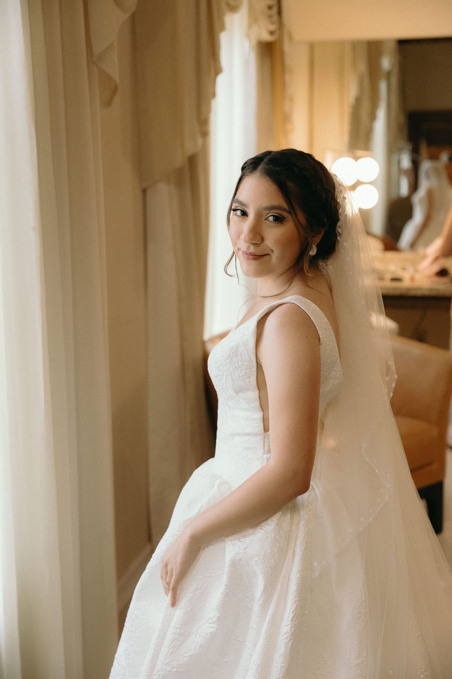 A bride in a classic white wedding gown poses elegantly by a sunlit window.