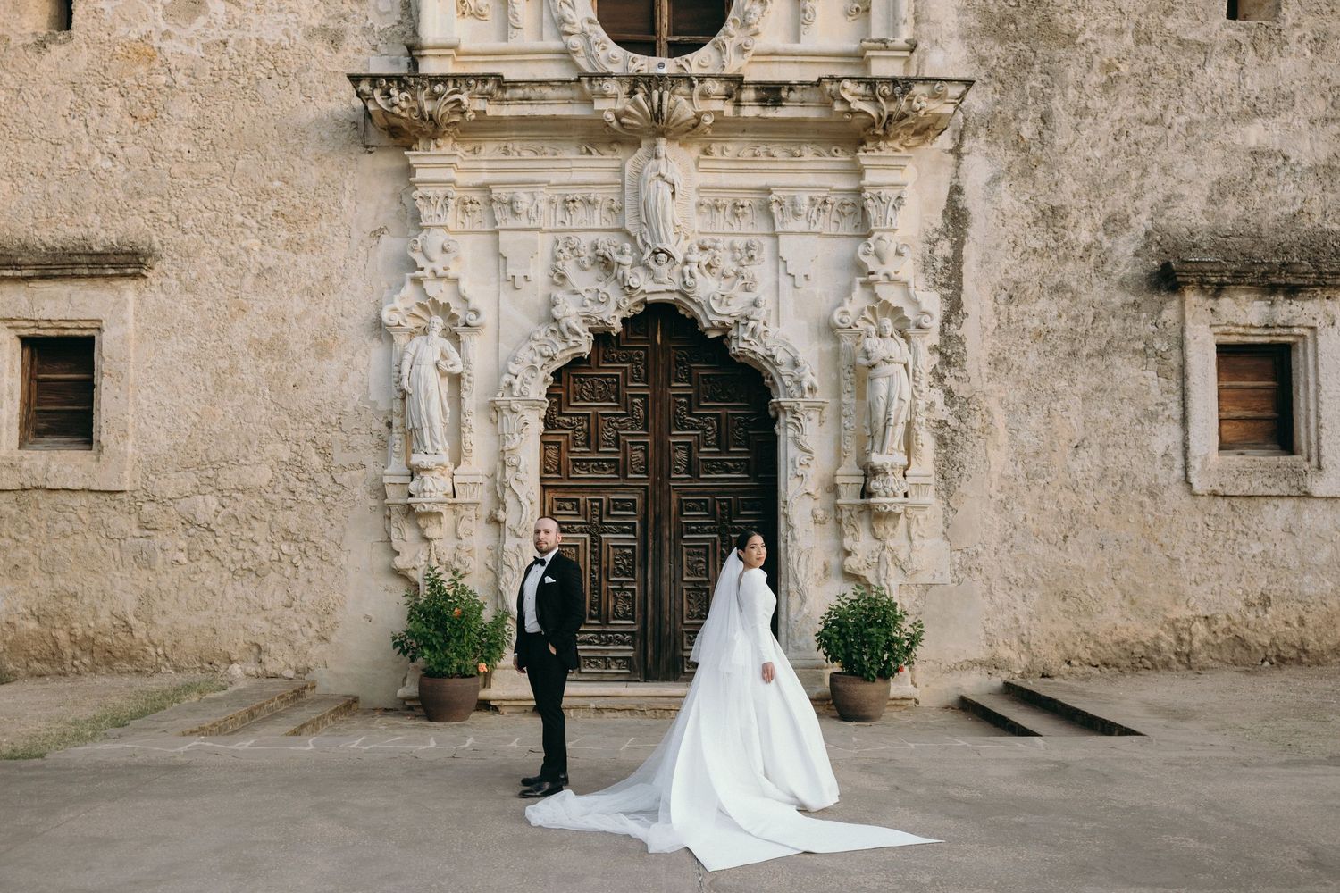 A bride and groom stand before an elaborate Spanish colonial church entrance with carved statues.