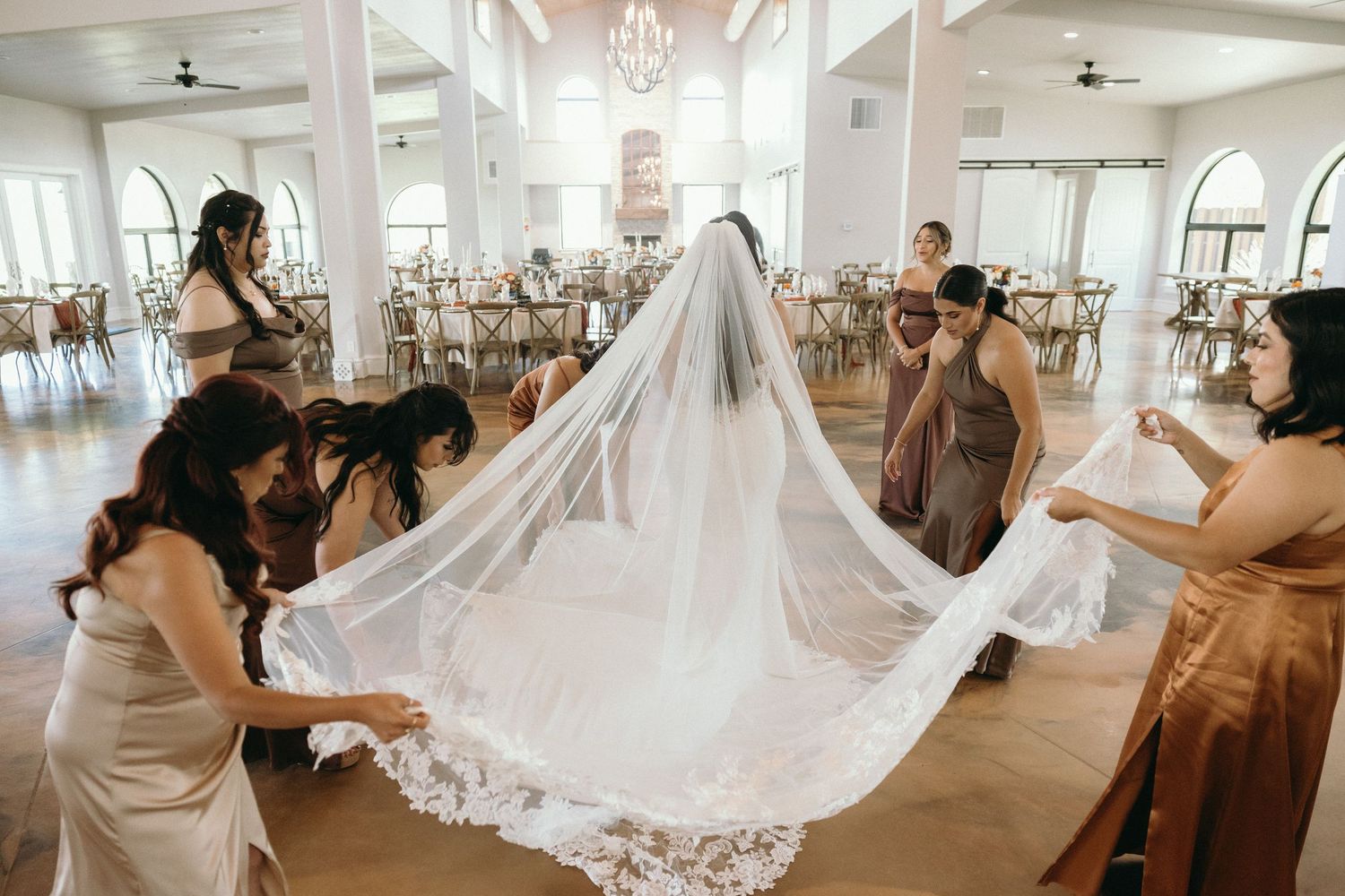 Wedding party members help adjust a flowing white wedding dress train in an elegant venue with chandeliers.