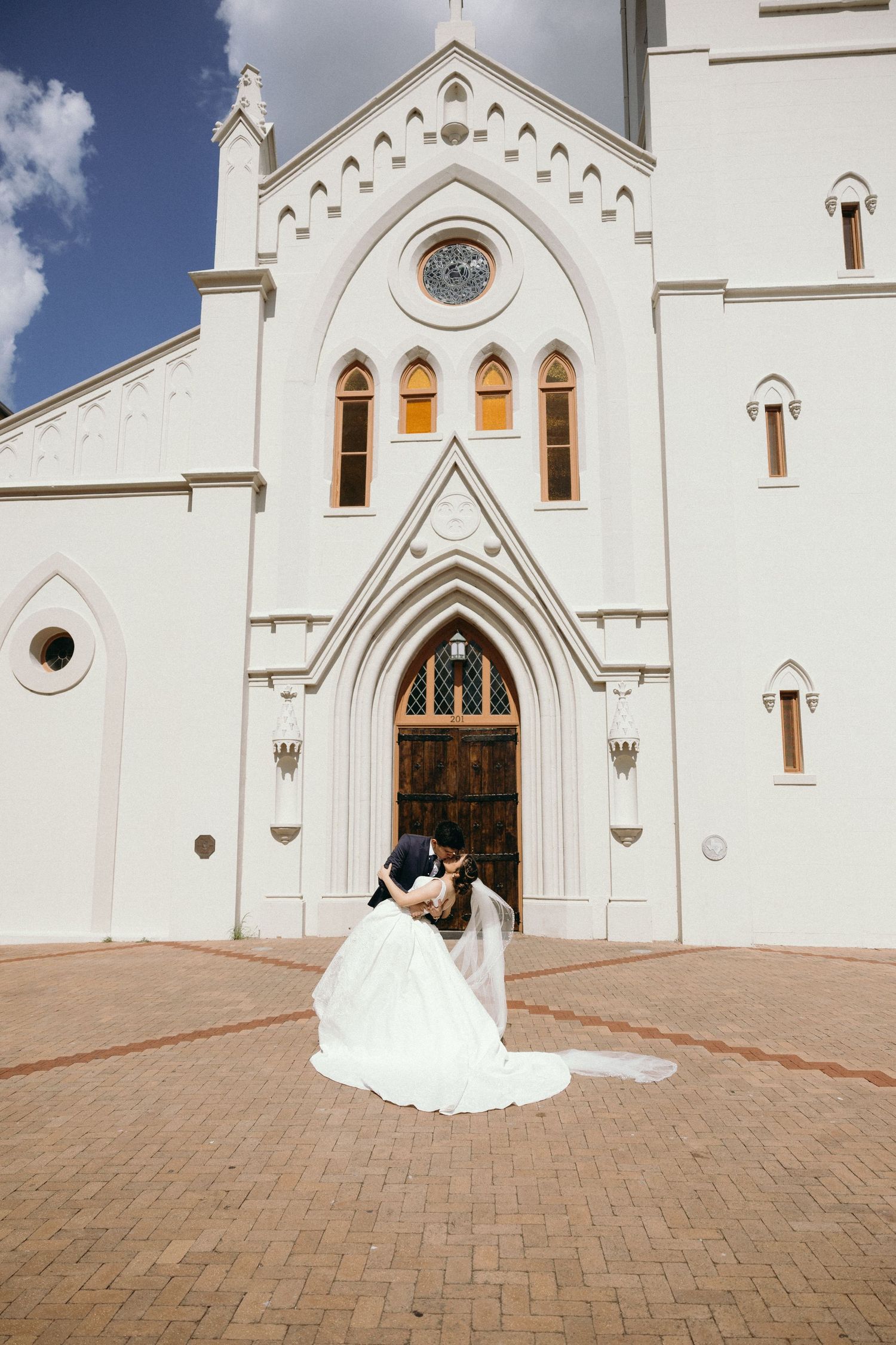 A wedding couple embraces in front of a grand white Gothic church entrance.