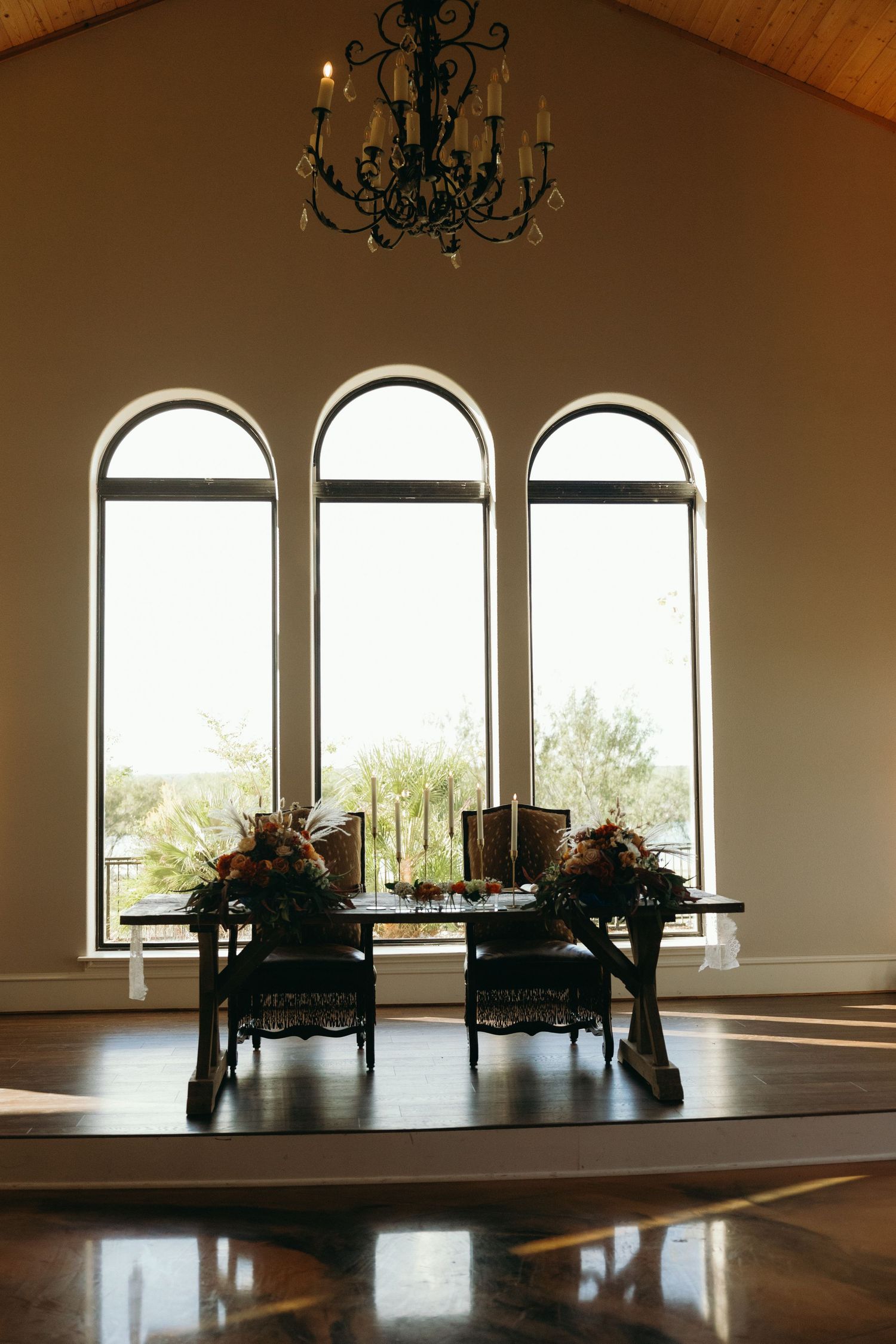 Elegant wedding reception setup with arched windows, chandelier, and decorated tables in a rustic venue.