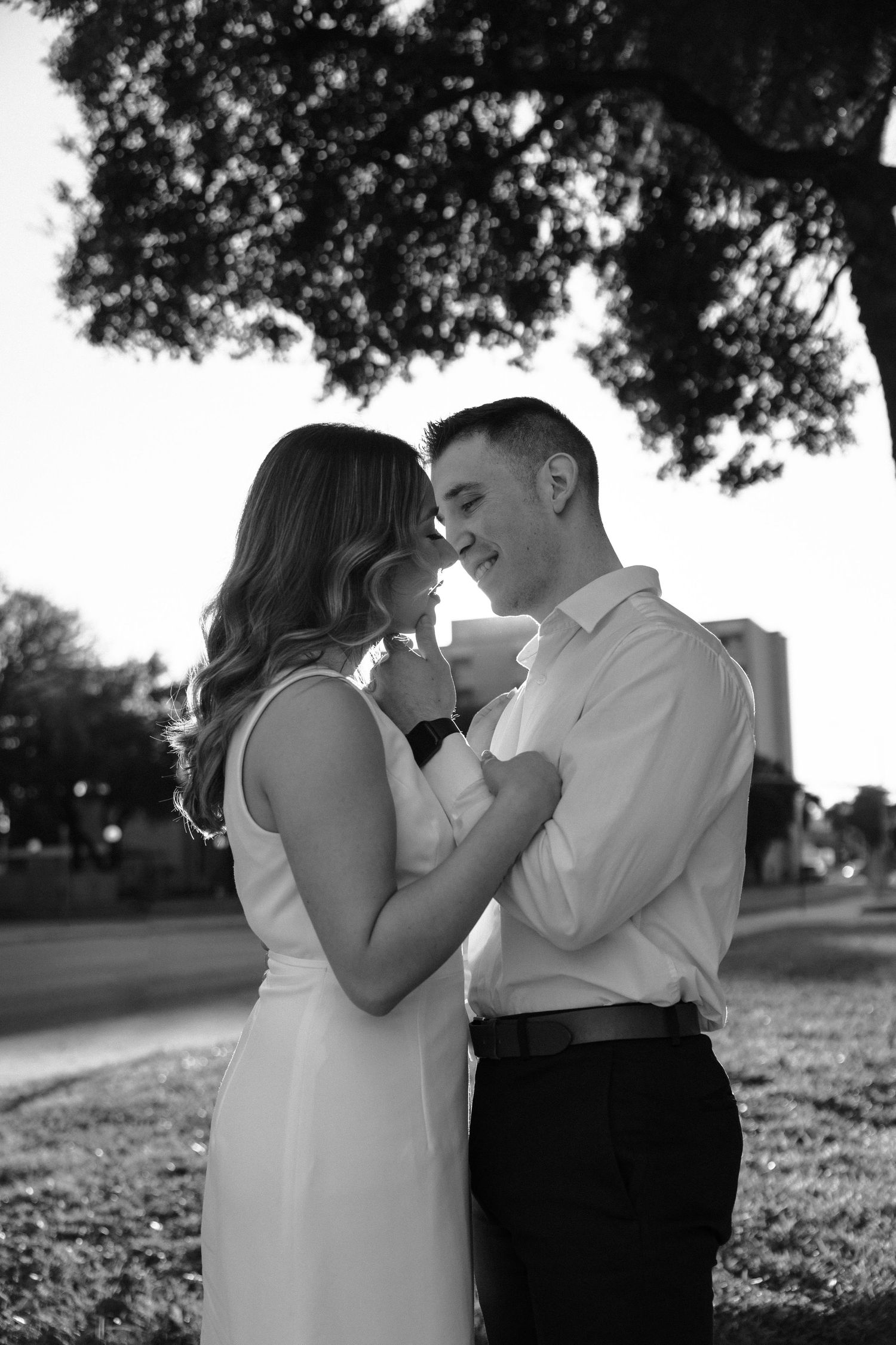 Black and white photo of a couple in formal attire embracing outdoors under trees.