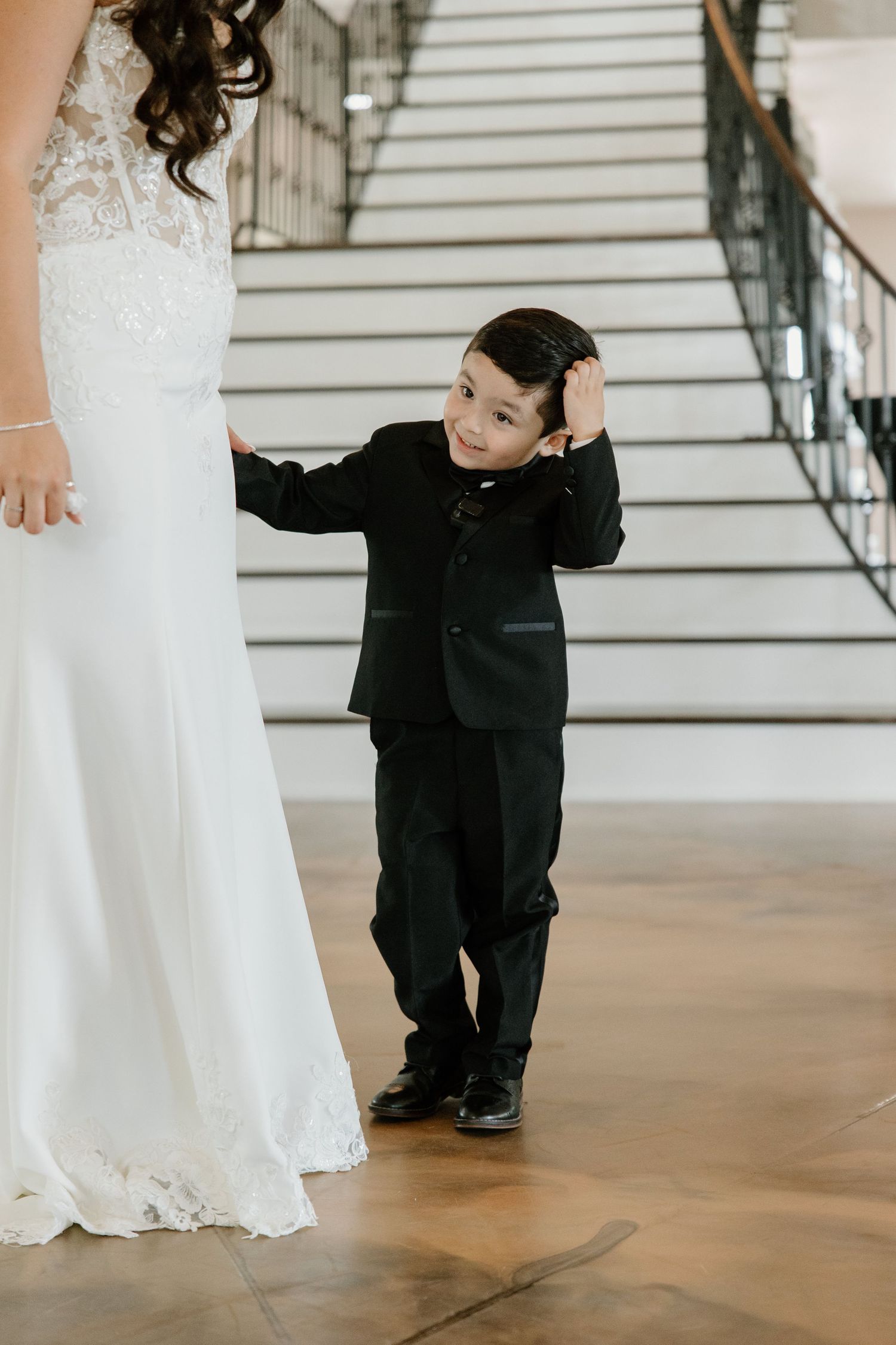 Young ring bearer in black suit kneels beside bride's white wedding dress.
