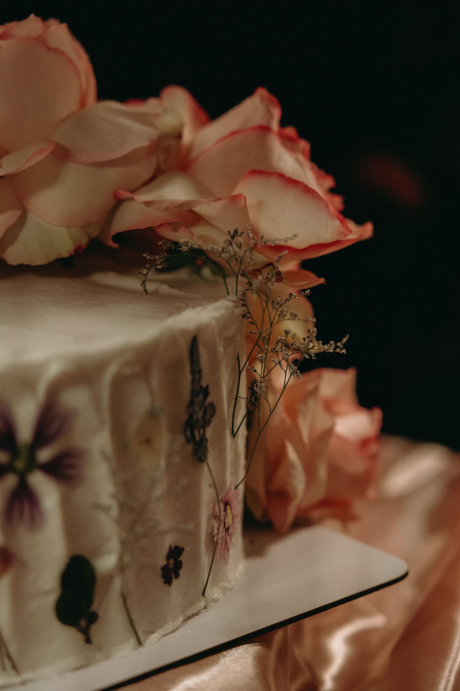 Close-up detail of a white wedding cake decorated with delicate pink roses against a dark background.