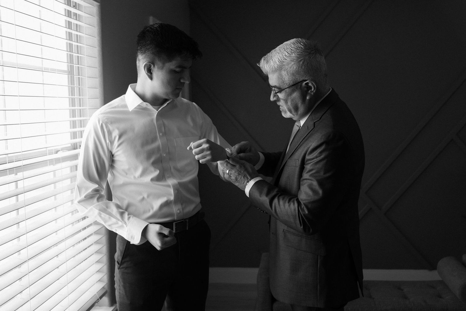 Black and white photograph showing two figures in conversation near a window with venetian blinds.