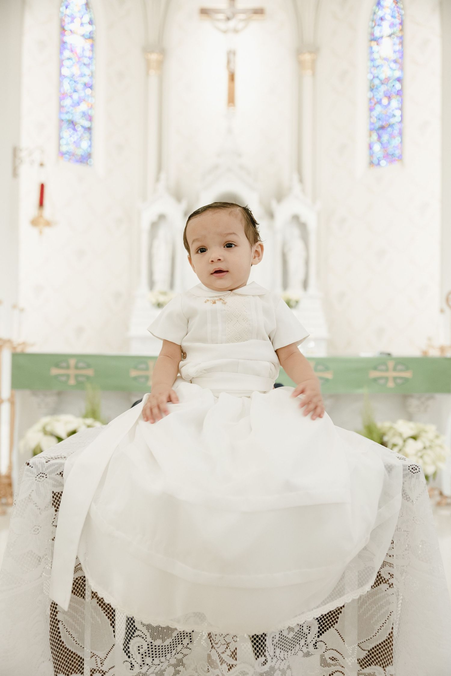 Baby in traditional white baptismal gown seated in church with stained glass windows and crucifix.