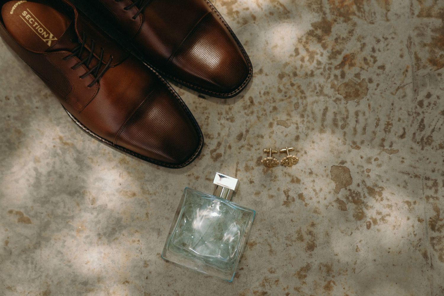 Brown leather dress shoes and cologne bottle arranged on rustic concrete surface.