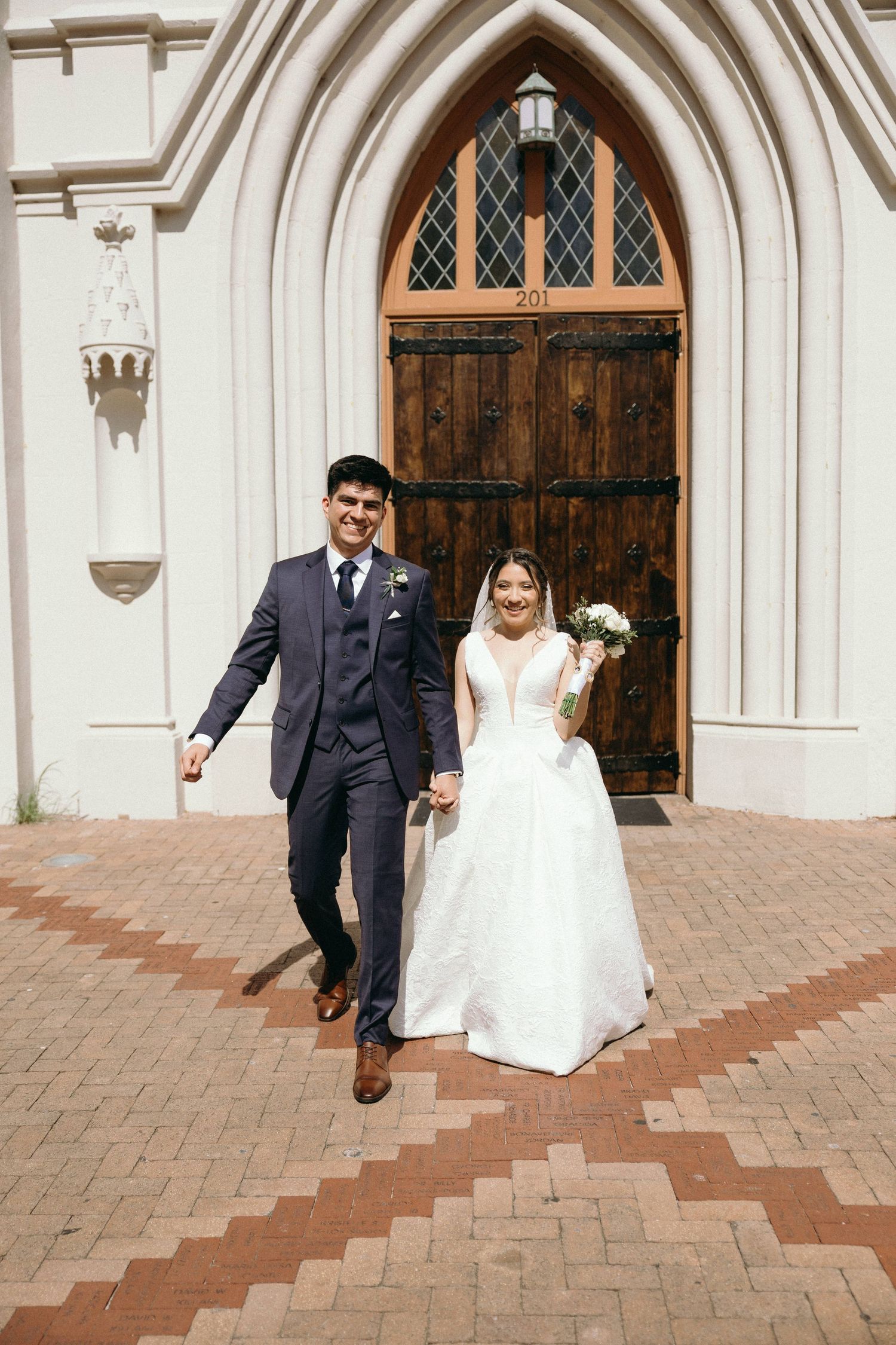 Couple walking down brick steps outside a Gothic church with arched wooden doors.
