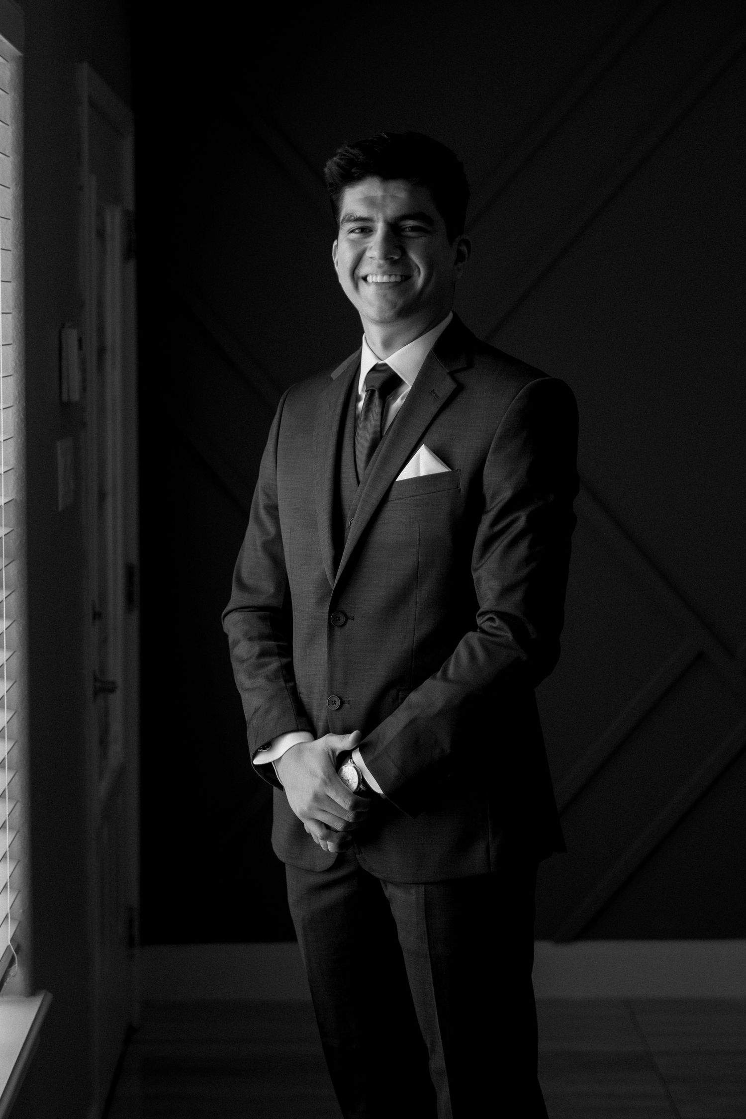 A formal portrait of a groom in a classic black suit standing by a window.