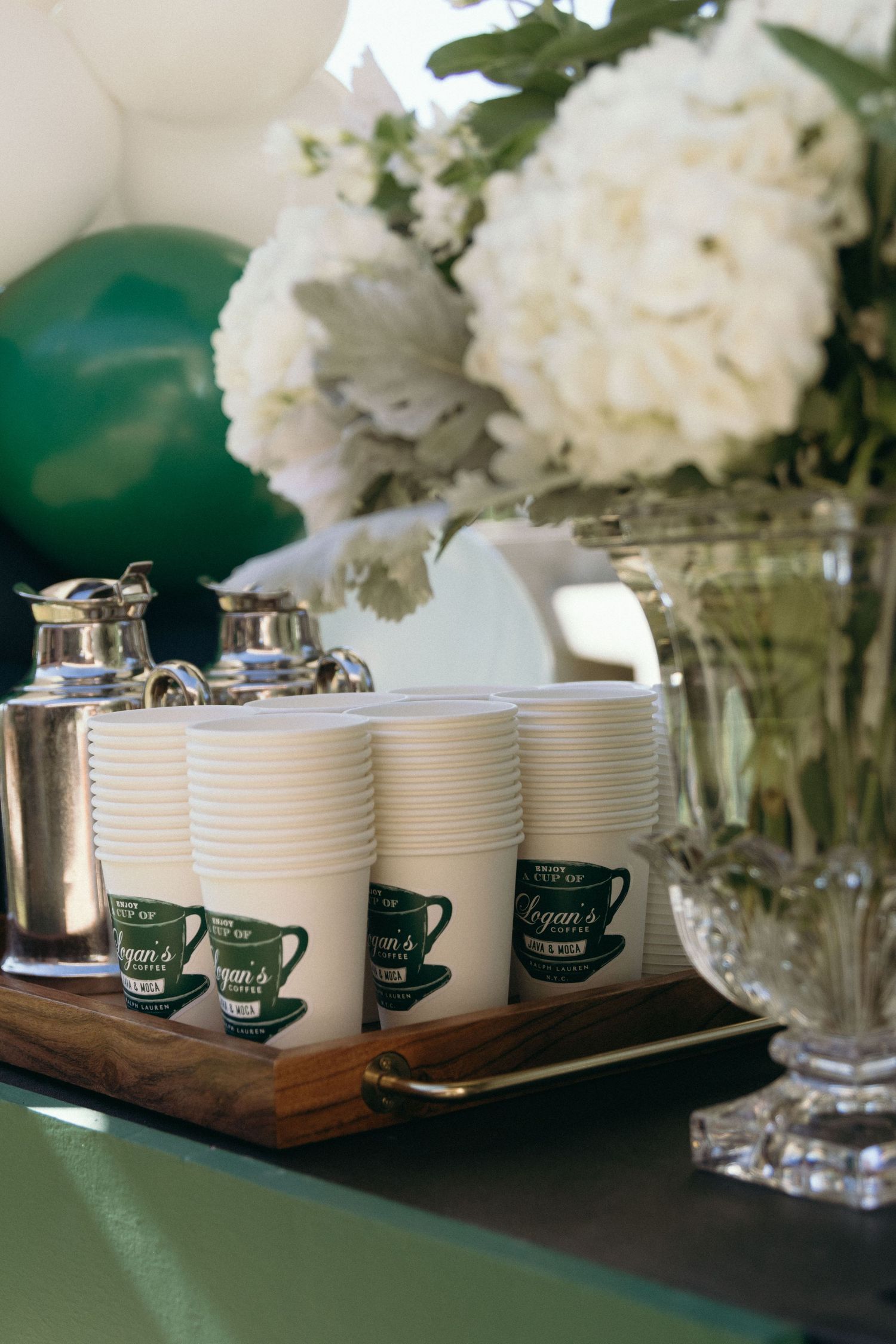 White coffee mugs arranged in a row with green tags on a dark wooden surface next to white floral arrangements.