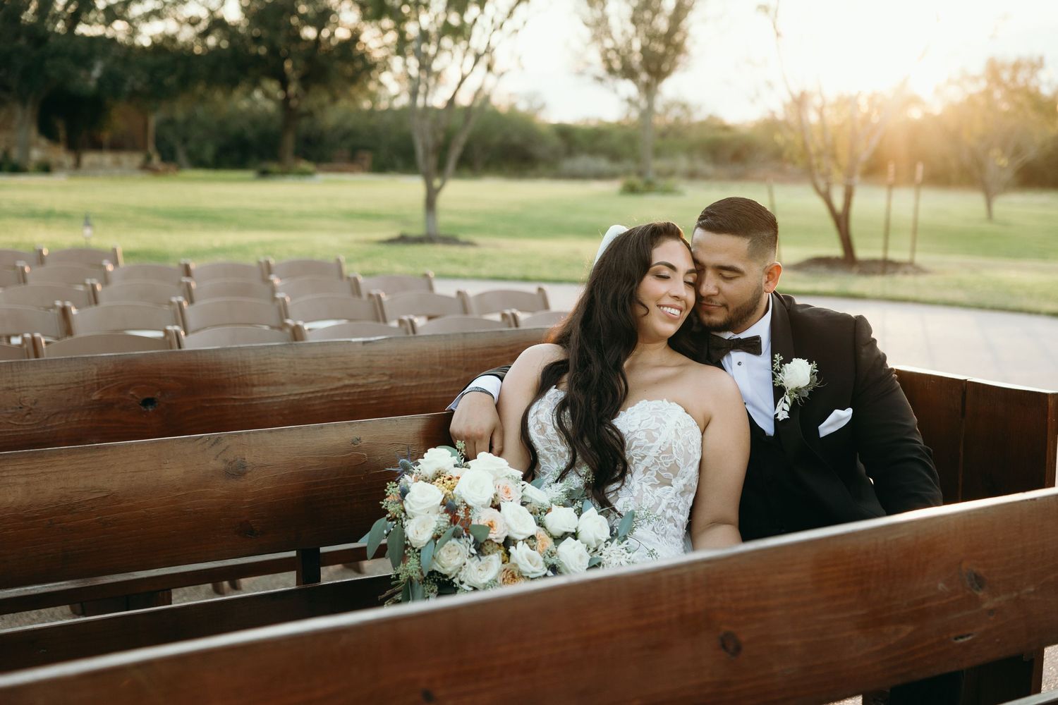 A couple sharing an intimate moment on wooden church pews outdoors with a white floral bouquet.