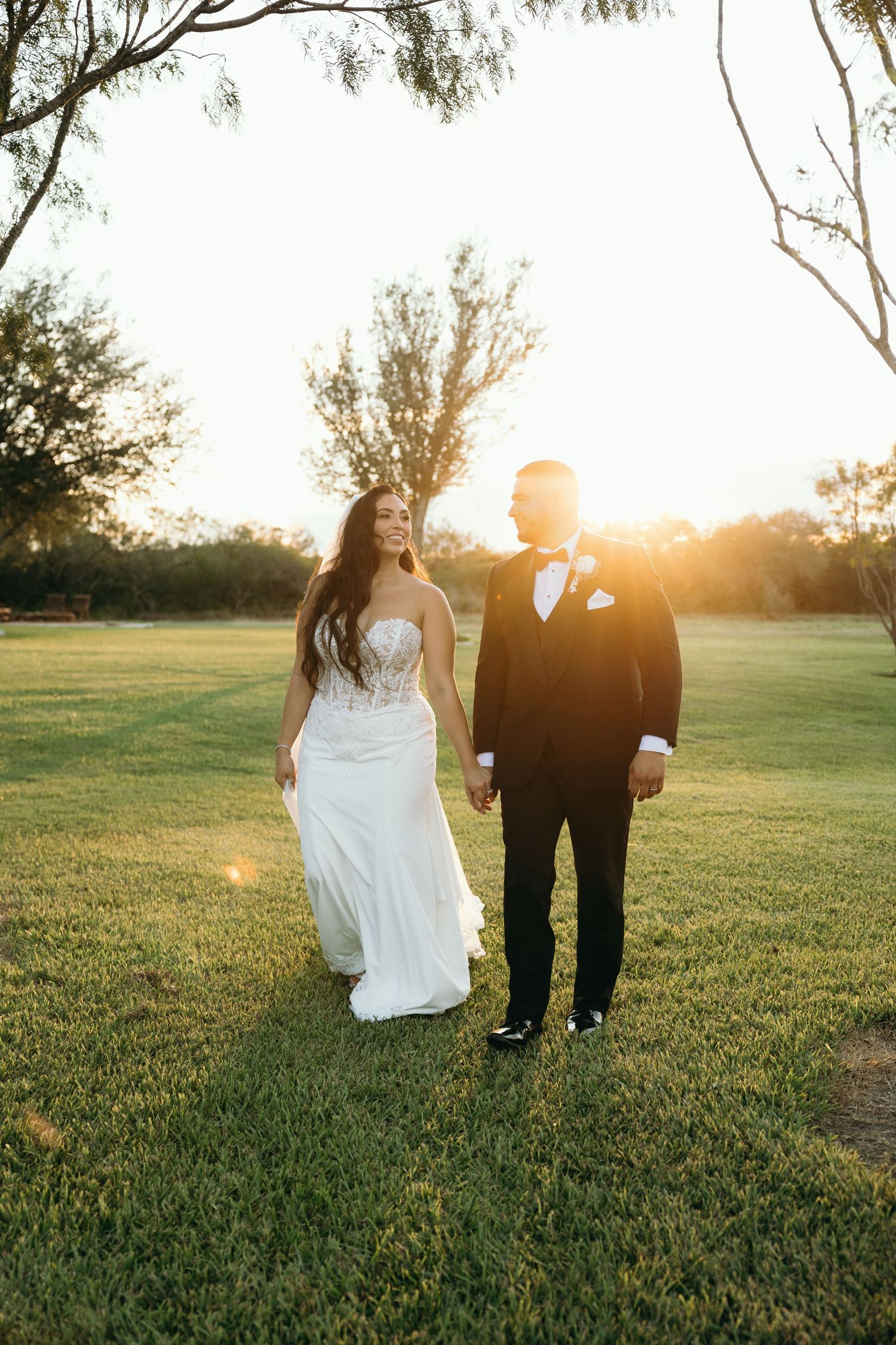 A wedding couple walking together on grass during golden hour sunset with trees in the background.