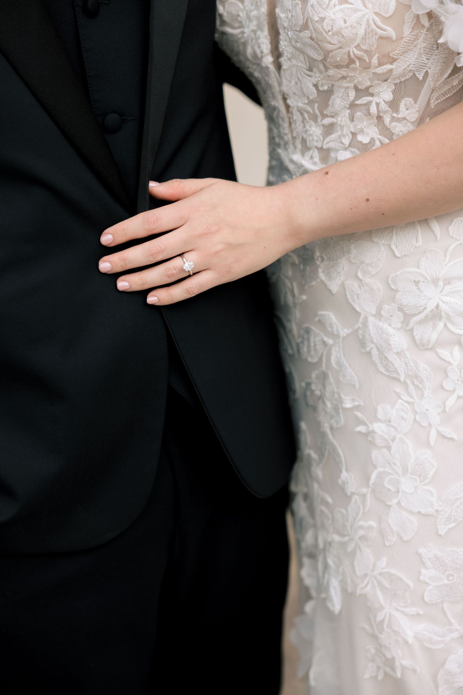 Bride in lace wedding gown rests hand on groom in black tuxedo, showcasing diamond engagement ring.