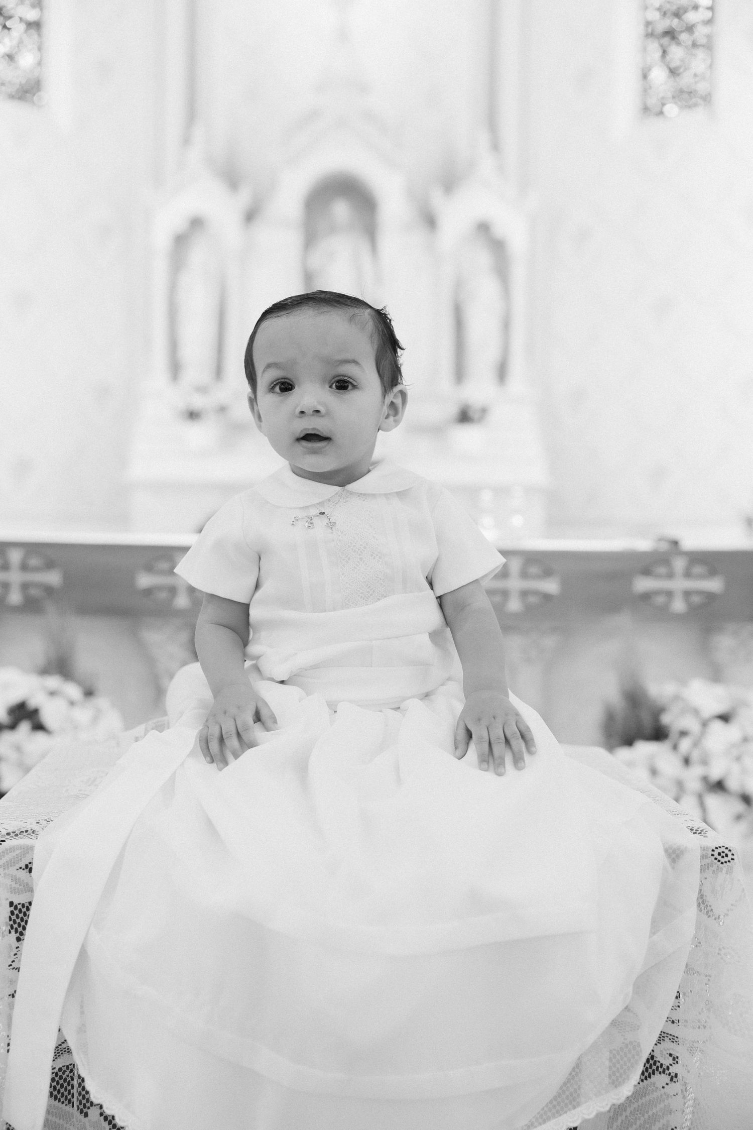 Small child wearing white christening gown seated in ornate church setting.