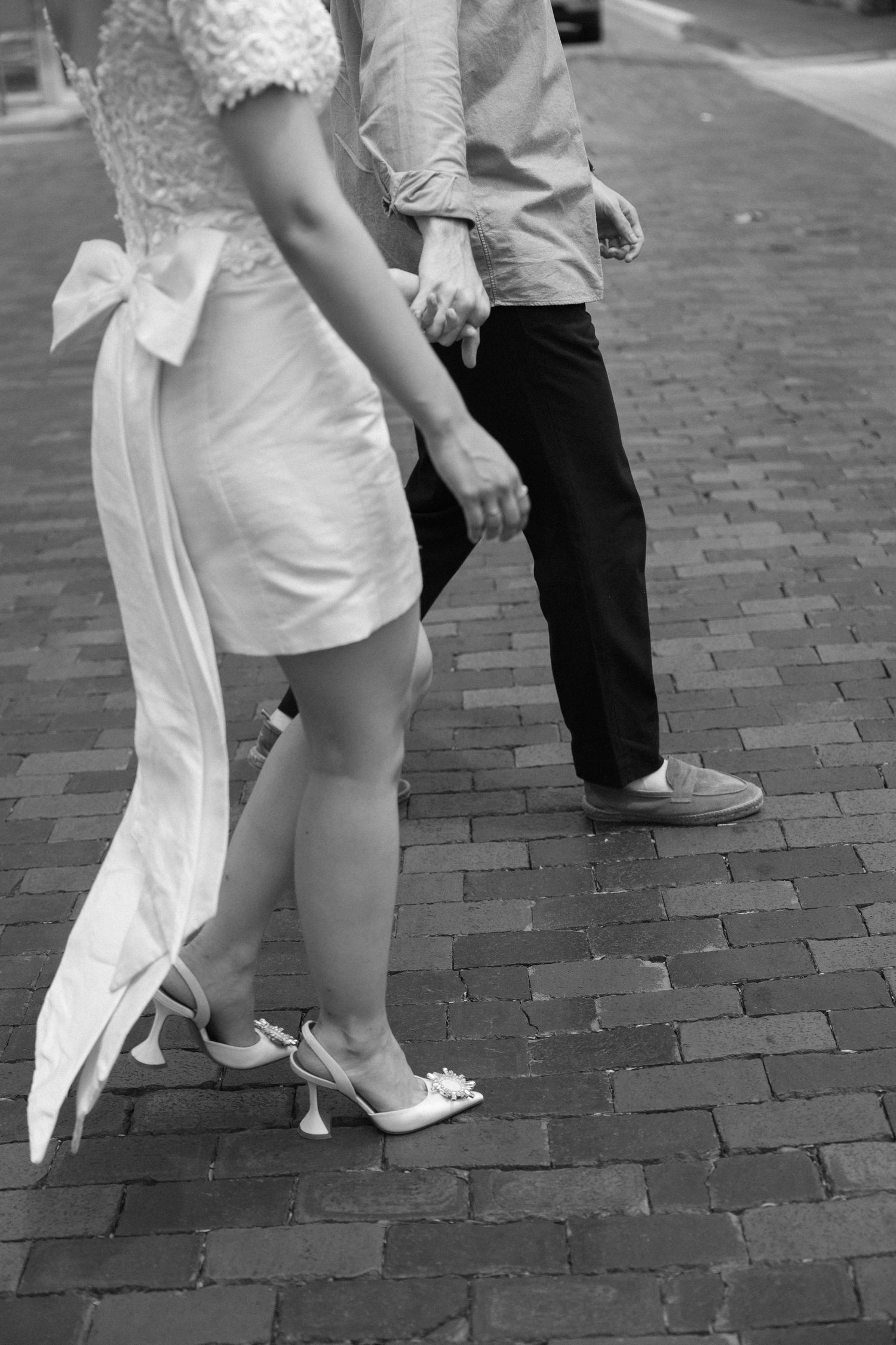 Black and white photo of formal shoes and dress hems on brick pavement.
