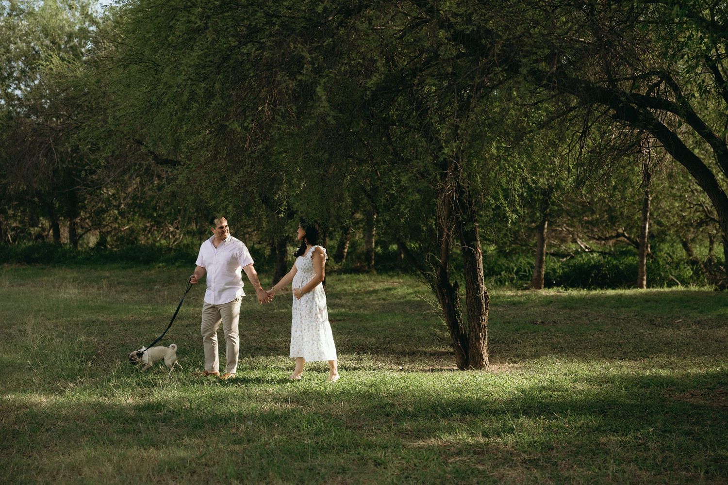A couple in white outfits walks their dog through a serene park setting.