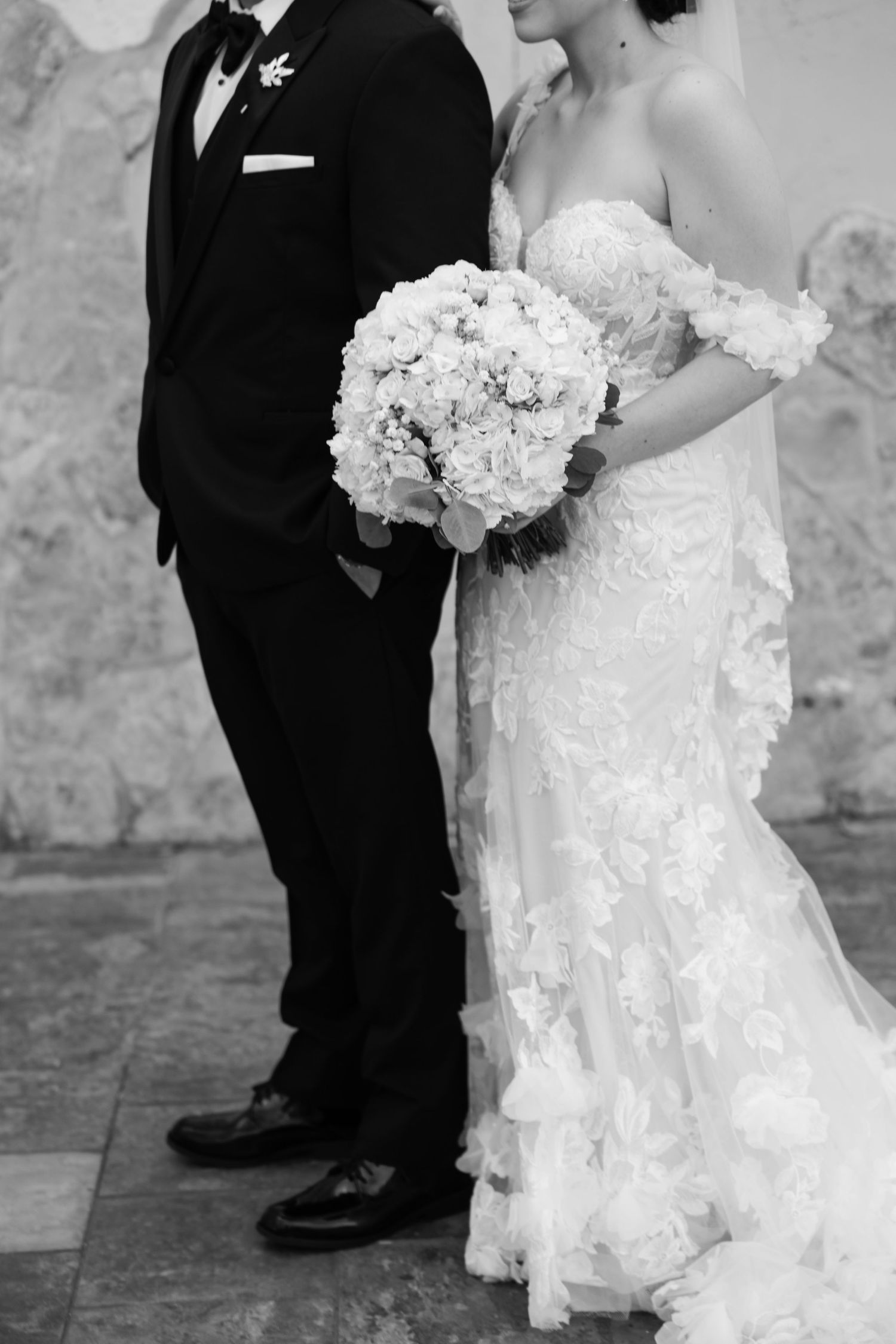 Black and white photo of bride in lace gown holding floral bouquet standing beside groom in tuxedo.