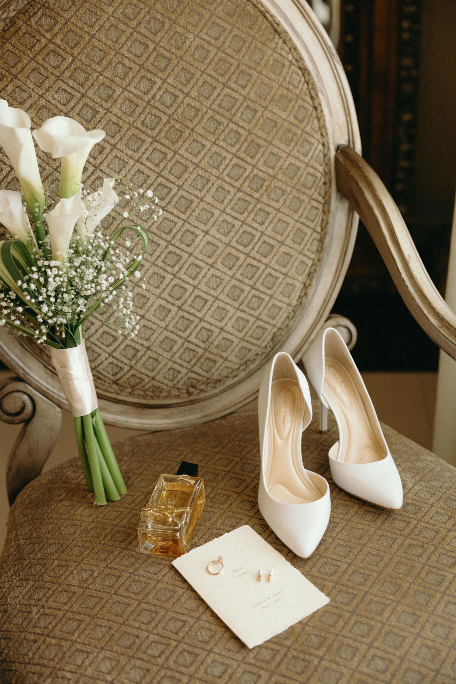 White bridal shoes and calla lily bouquet arranged on an ornate chair with geometric pattern.