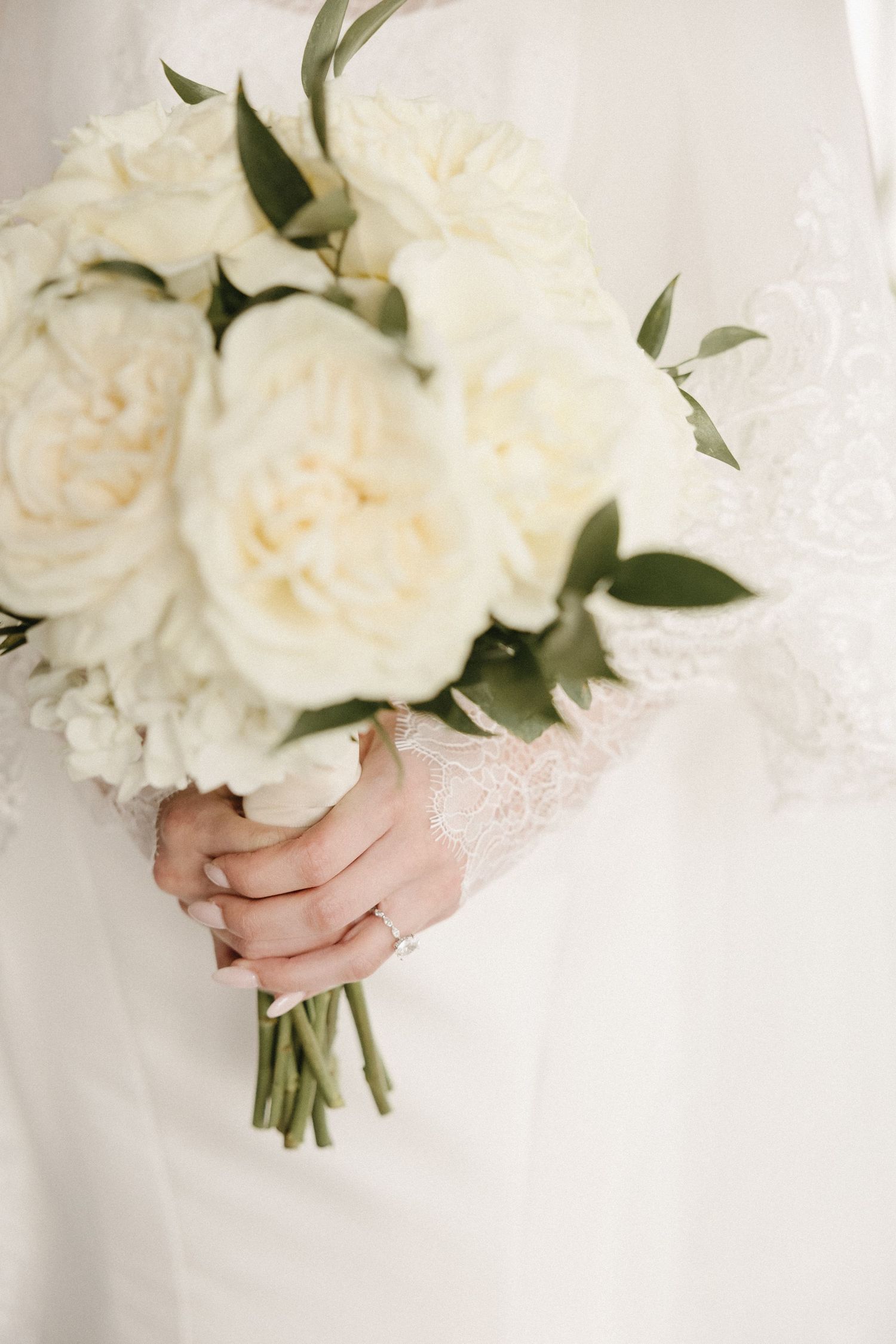 Classic white rose bridal bouquet with green foliage held against lace dress.