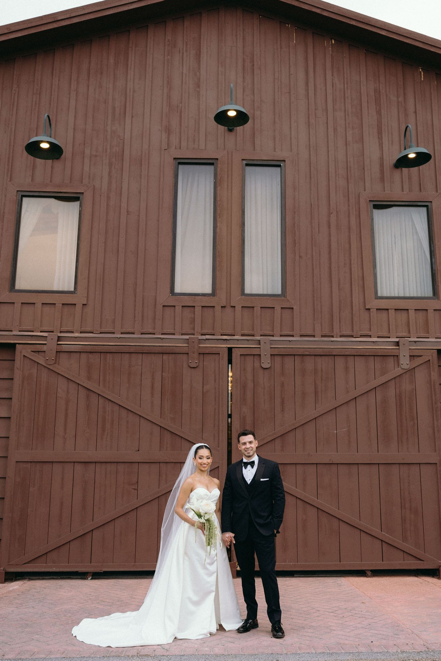 Wedding couple poses in front of a rustic brown barn with large wooden doors.