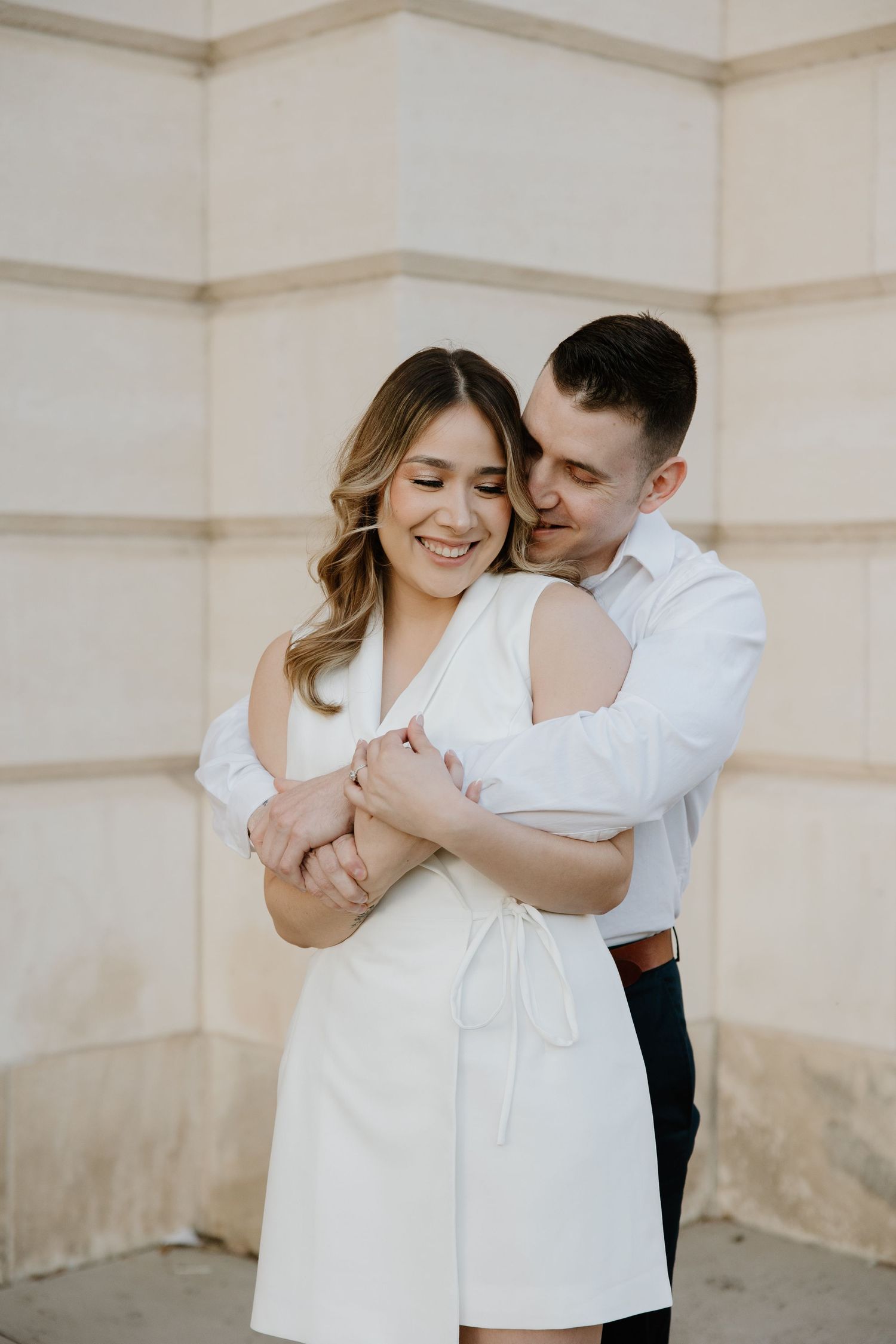 Romantic couple in white clothing embracing against a neutral wall background.