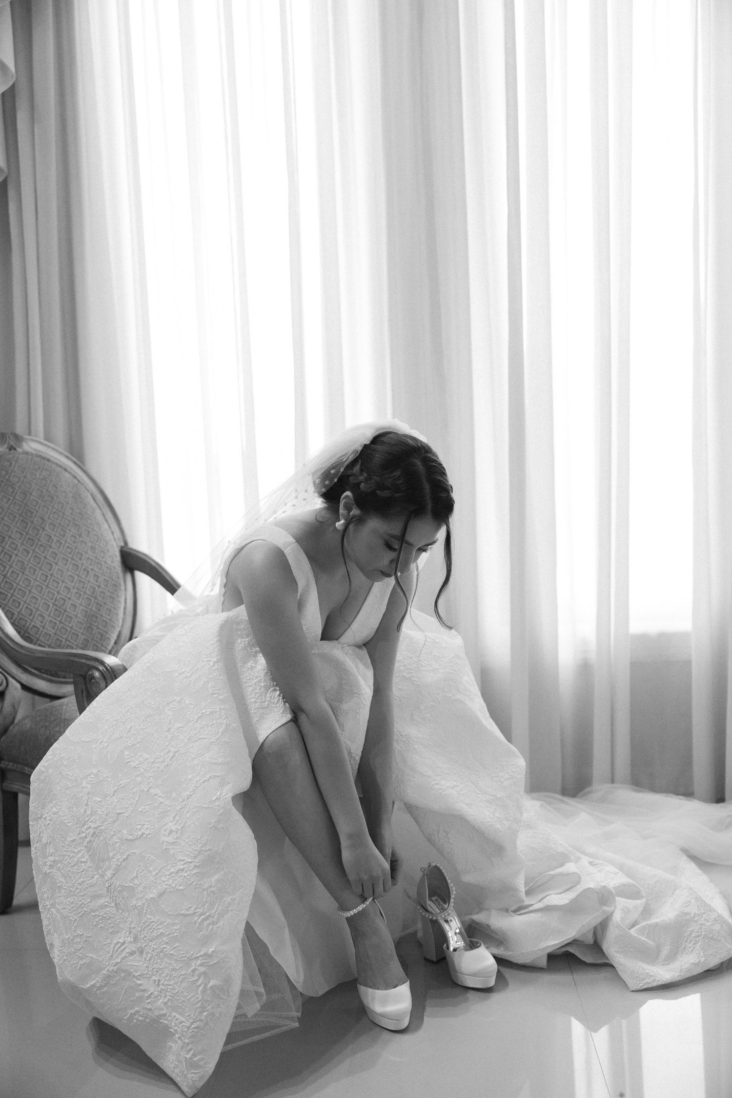 A bride in white adjusts her shoe while sitting in an elegant room.