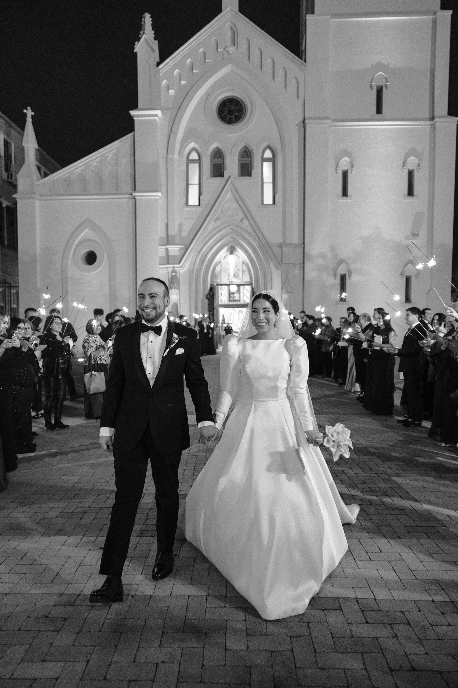 Wedding party exits Gothic church at night with illuminated facade.