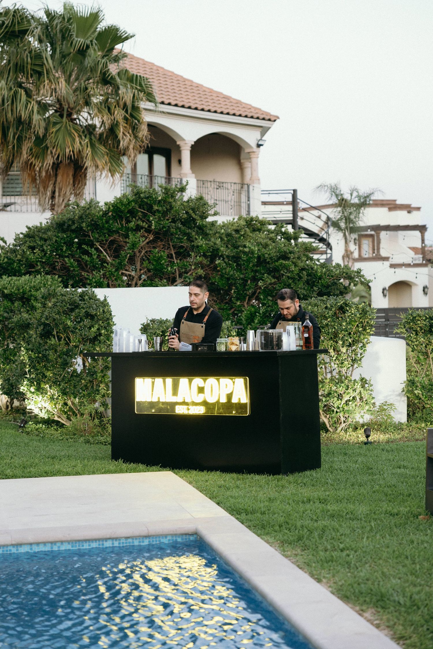 Illuminated DJ booth setup with 'MALACOPA' sign near a pool at an outdoor event.