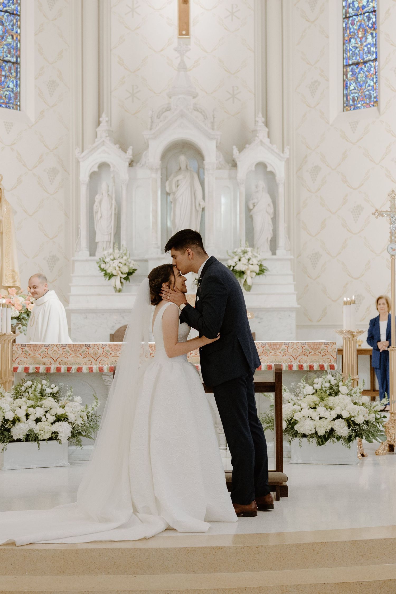 Wedding ceremony in a white church interior with floral arrangements and stained glass windows.