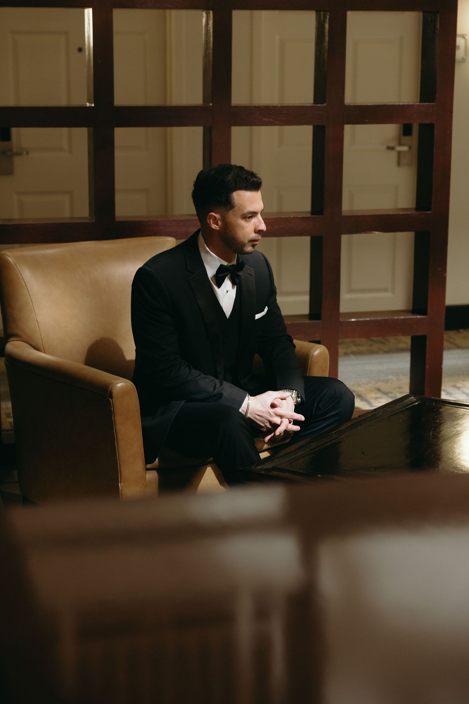 Man in tuxedo sits thoughtfully in elegant indoor setting.