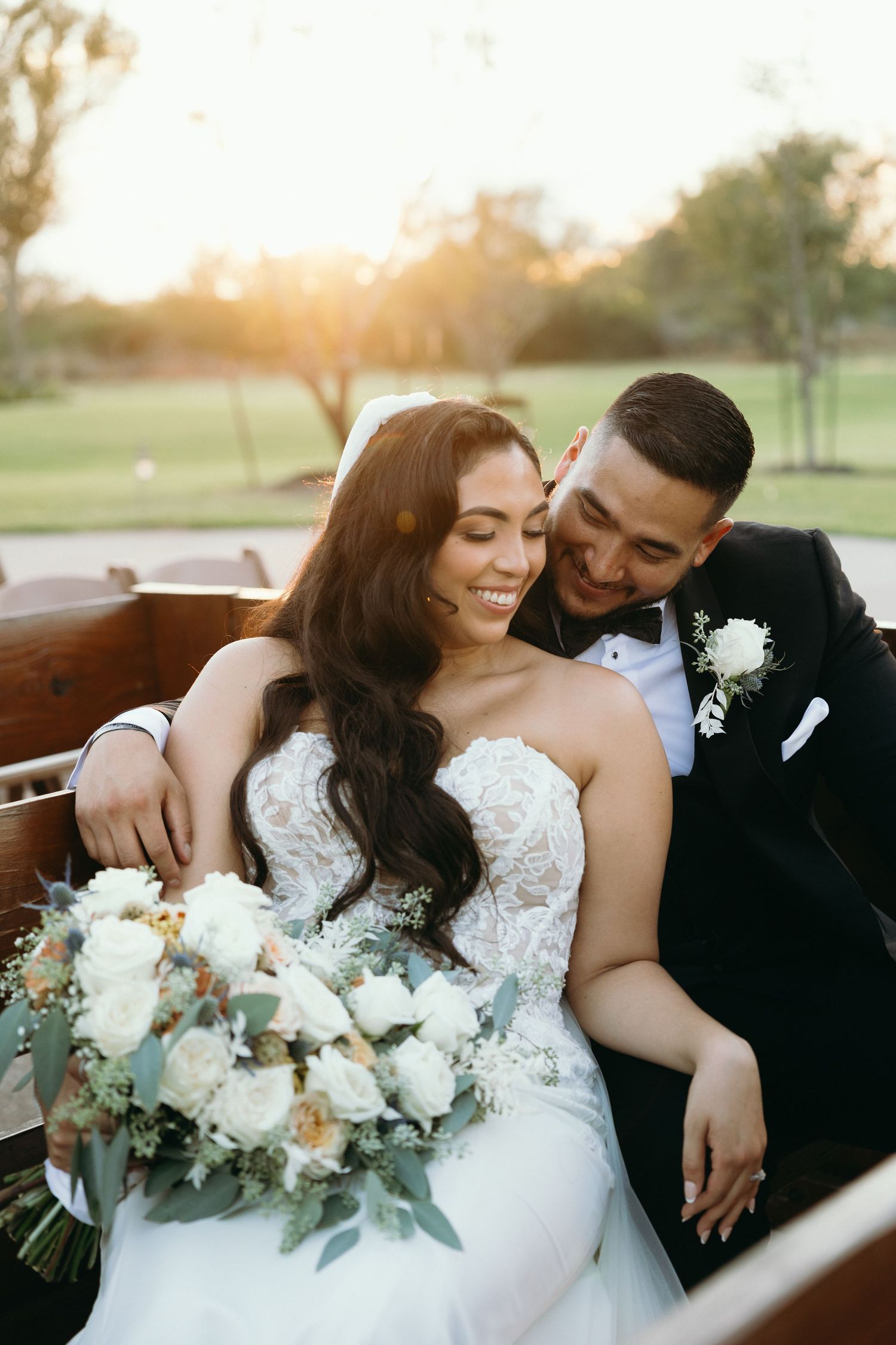 A couple sharing a joyful moment on wooden benches with a white and green floral bouquet at sunset.