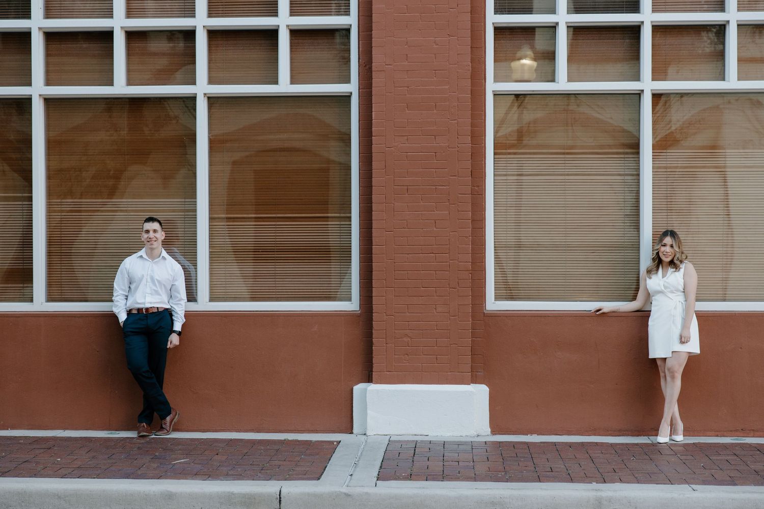 Couple in white outfits posing against a modern brick building with large windows.