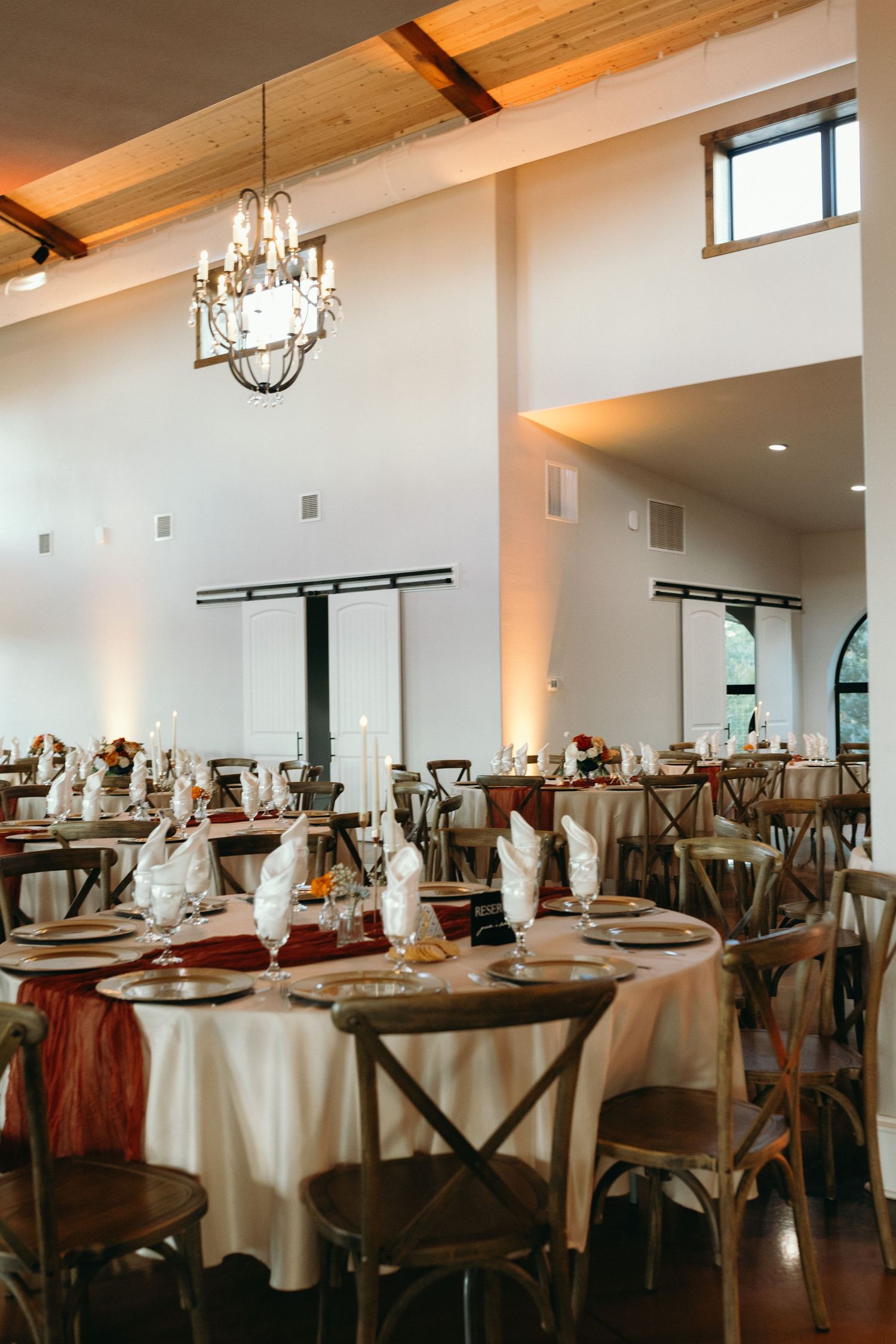 An elegant wedding reception setup with crystal chandeliers and round tables with crossback chairs.