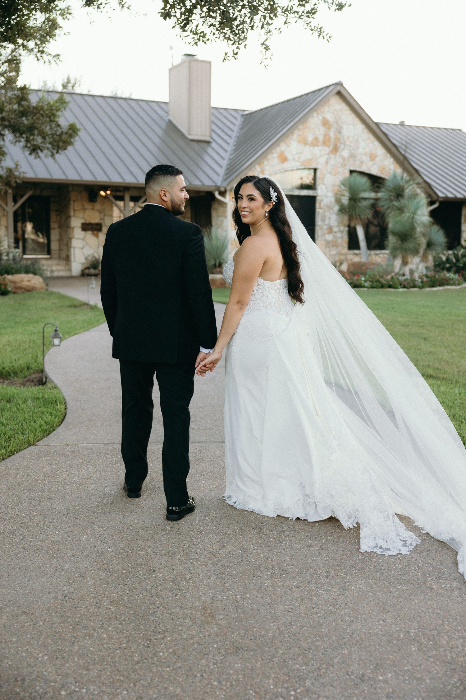 A newlywed couple walking hand in hand along a pathway in front of a stone building with a metal roof.