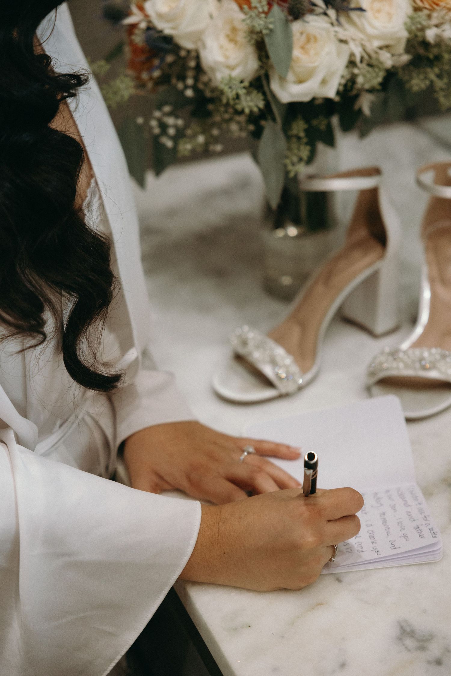 Close-up of hands writing wedding vows with floral arrangements and bridal shoes in background.