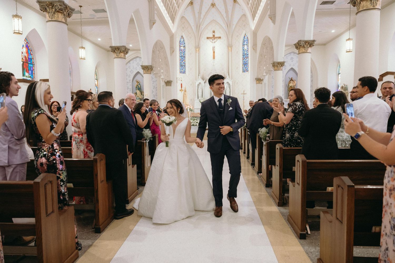 Wedding ceremony taking place in a beautiful white Gothic church with vaulted ceilings and wooden pews.