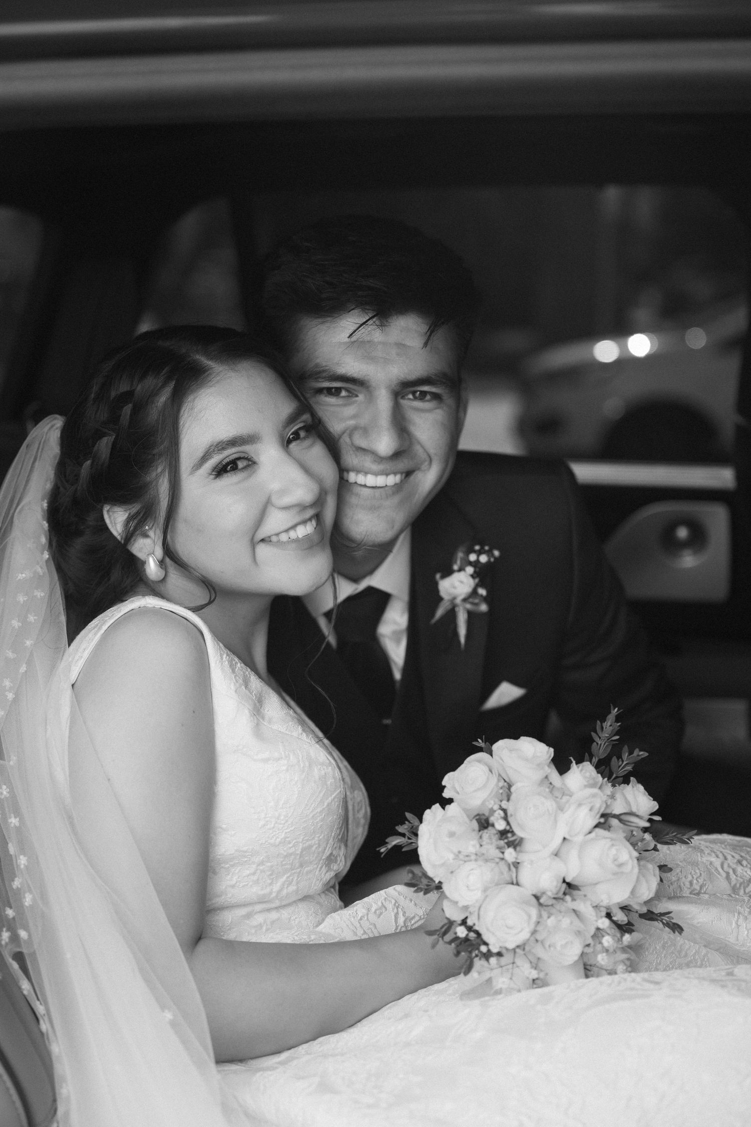 Newlyweds share intimate moments in a car with white floral bouquet and formal wedding attire.