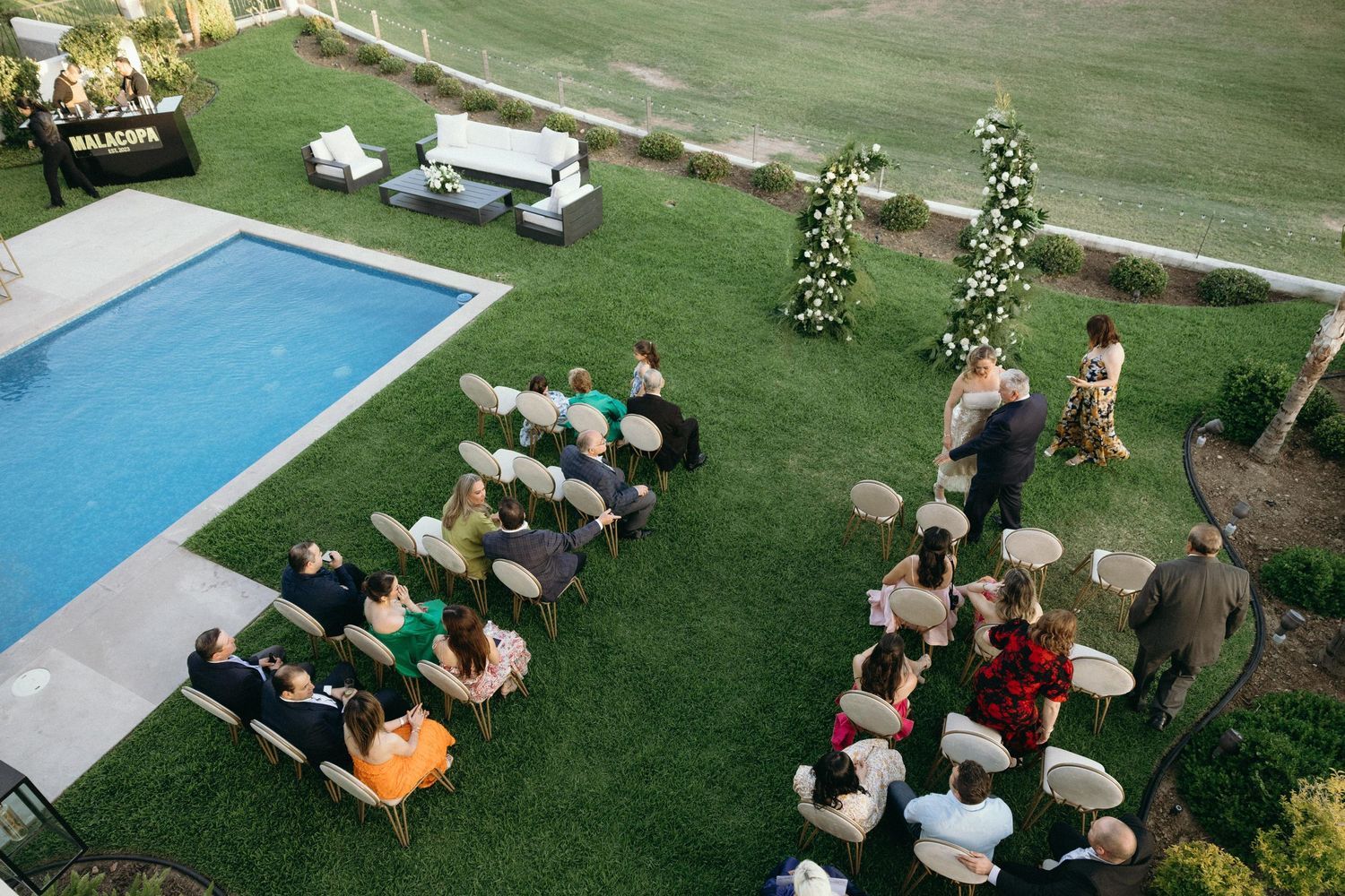 Aerial view of an outdoor wedding ceremony setup with chairs and guests on a lawn near a pool.