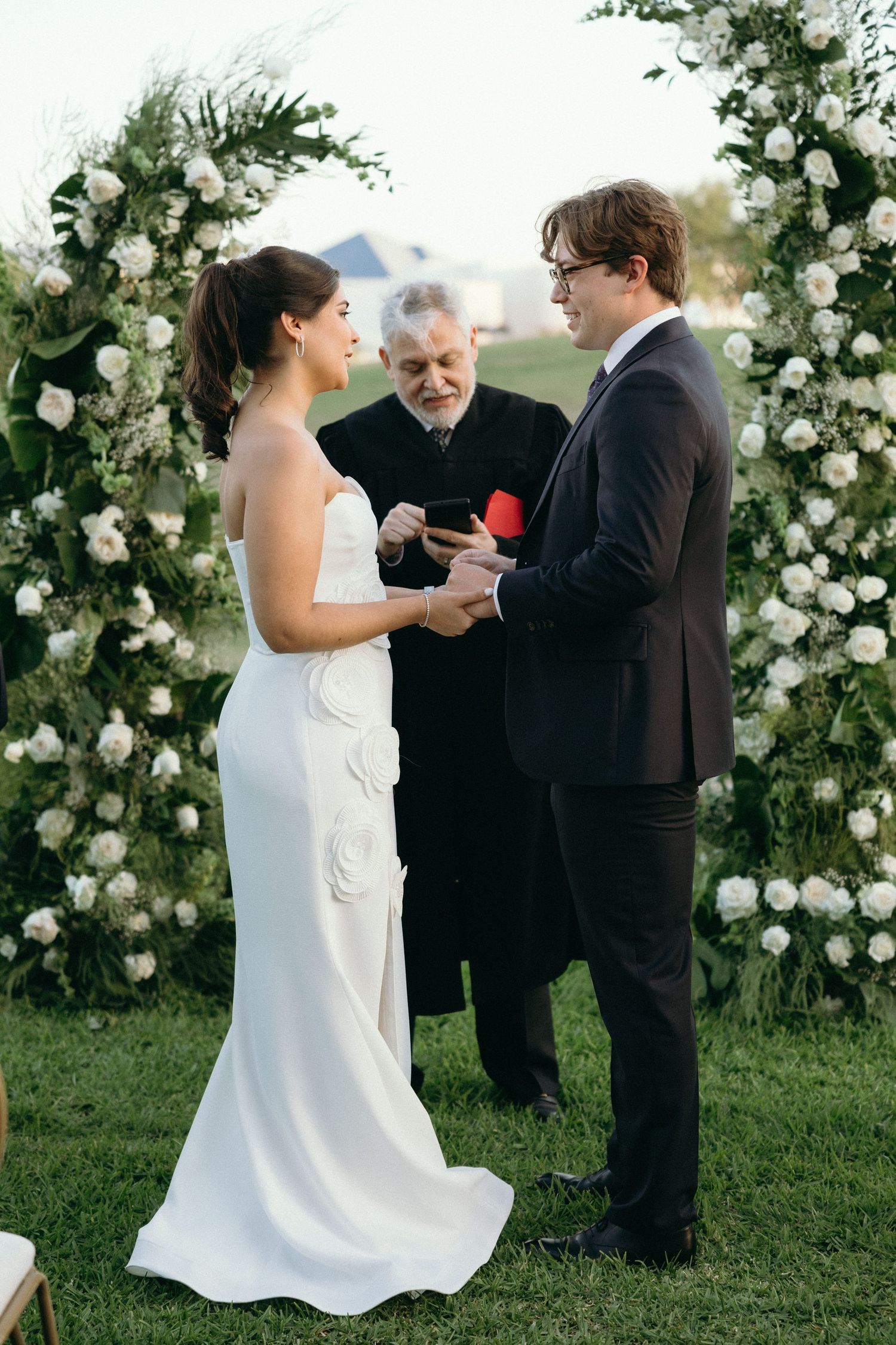 Outdoor wedding ceremony with white floral arch and guests gathered on a grassy lawn.