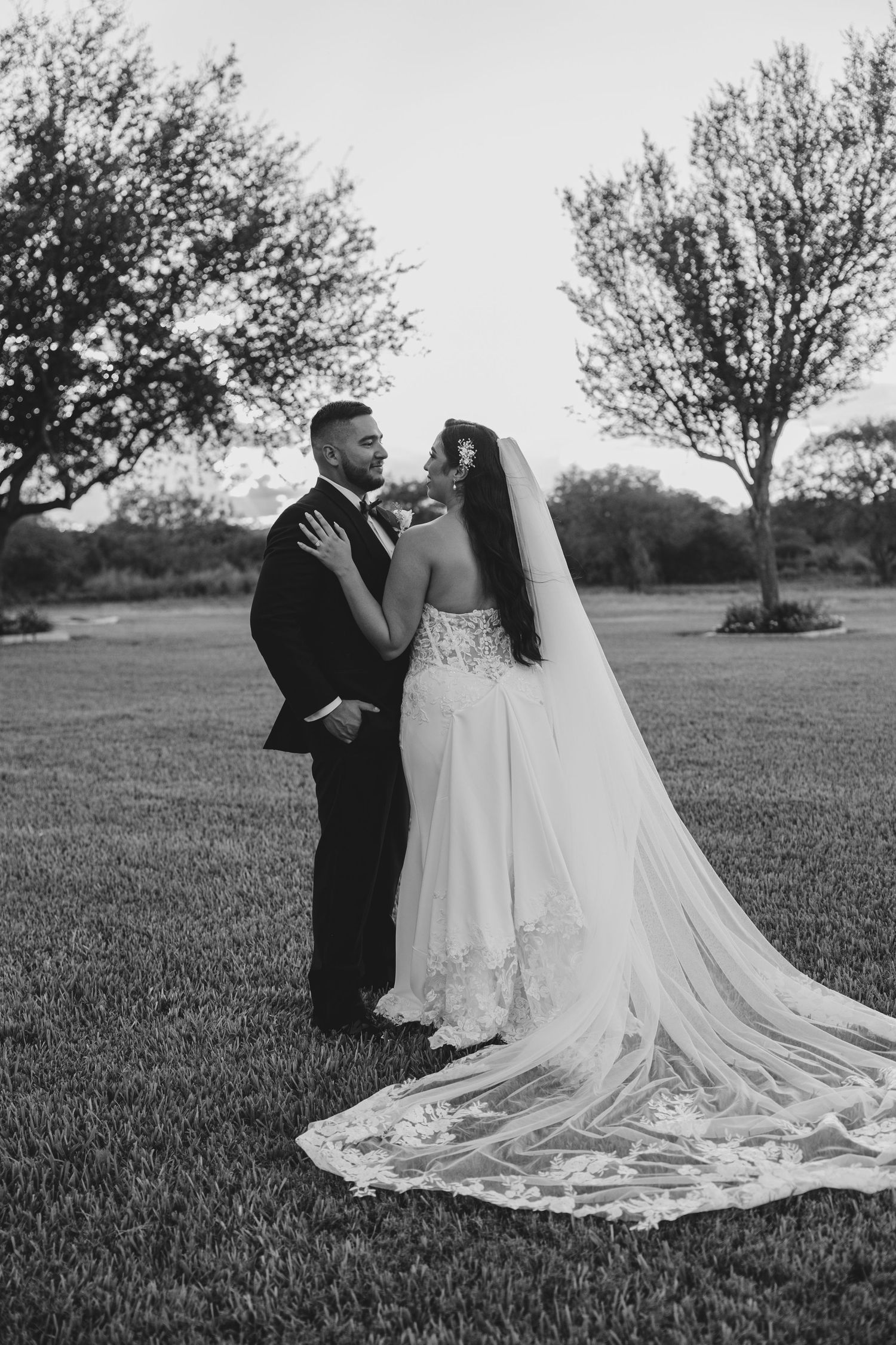 A romantic black and white wedding portrait of a couple embracing on a grassy lawn with trees in the background.