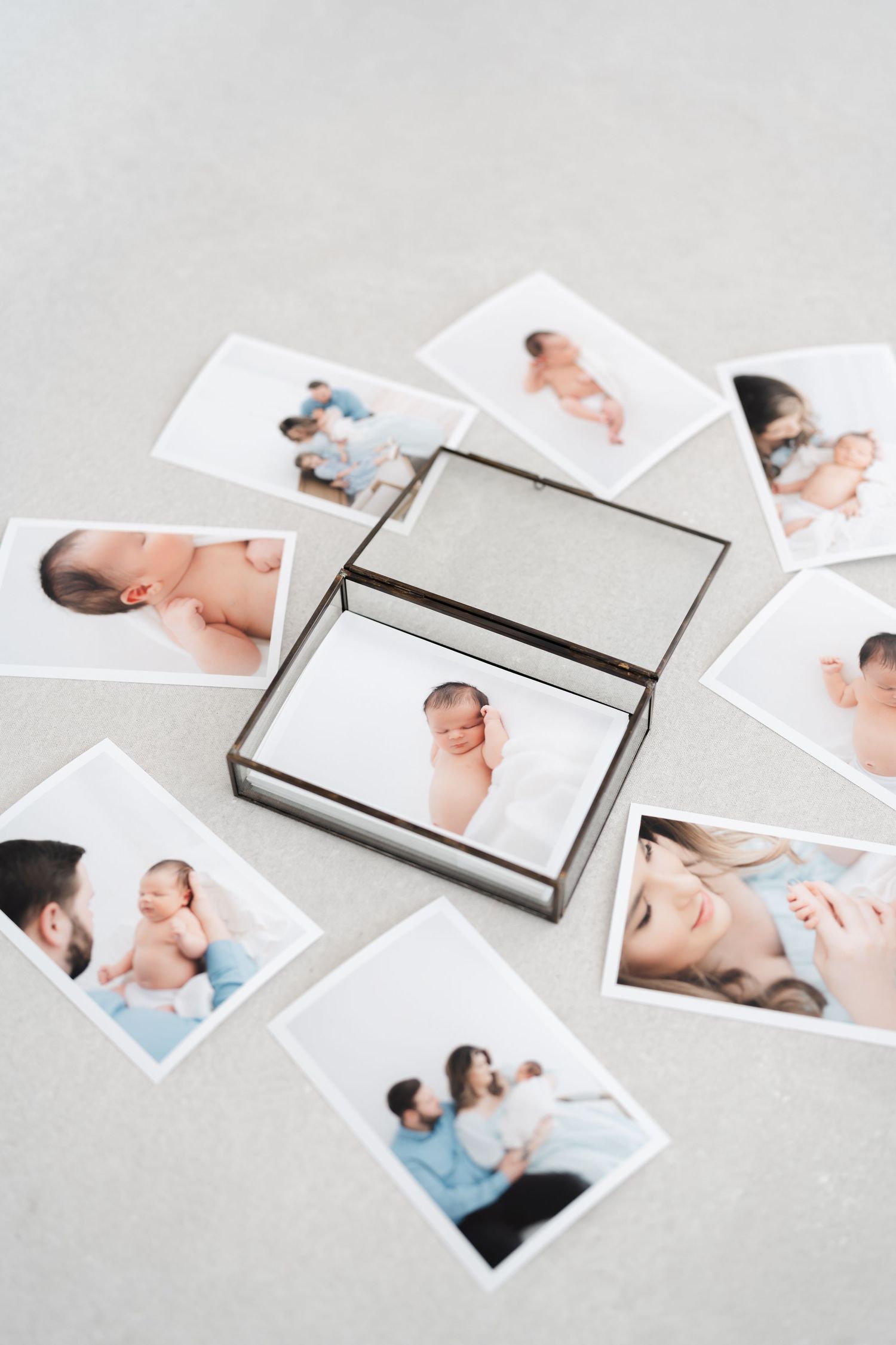 An open glass keepsake box surrounded by scattered newborn baby photos with family portraits on a light gray surface.