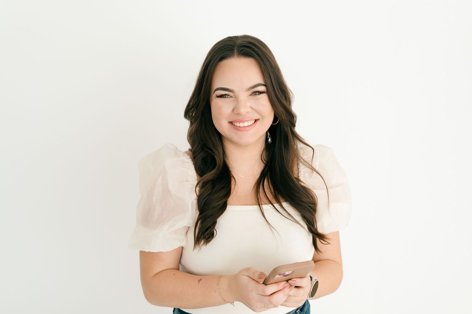 Smiling brunette woman in white top holding phone against white background.