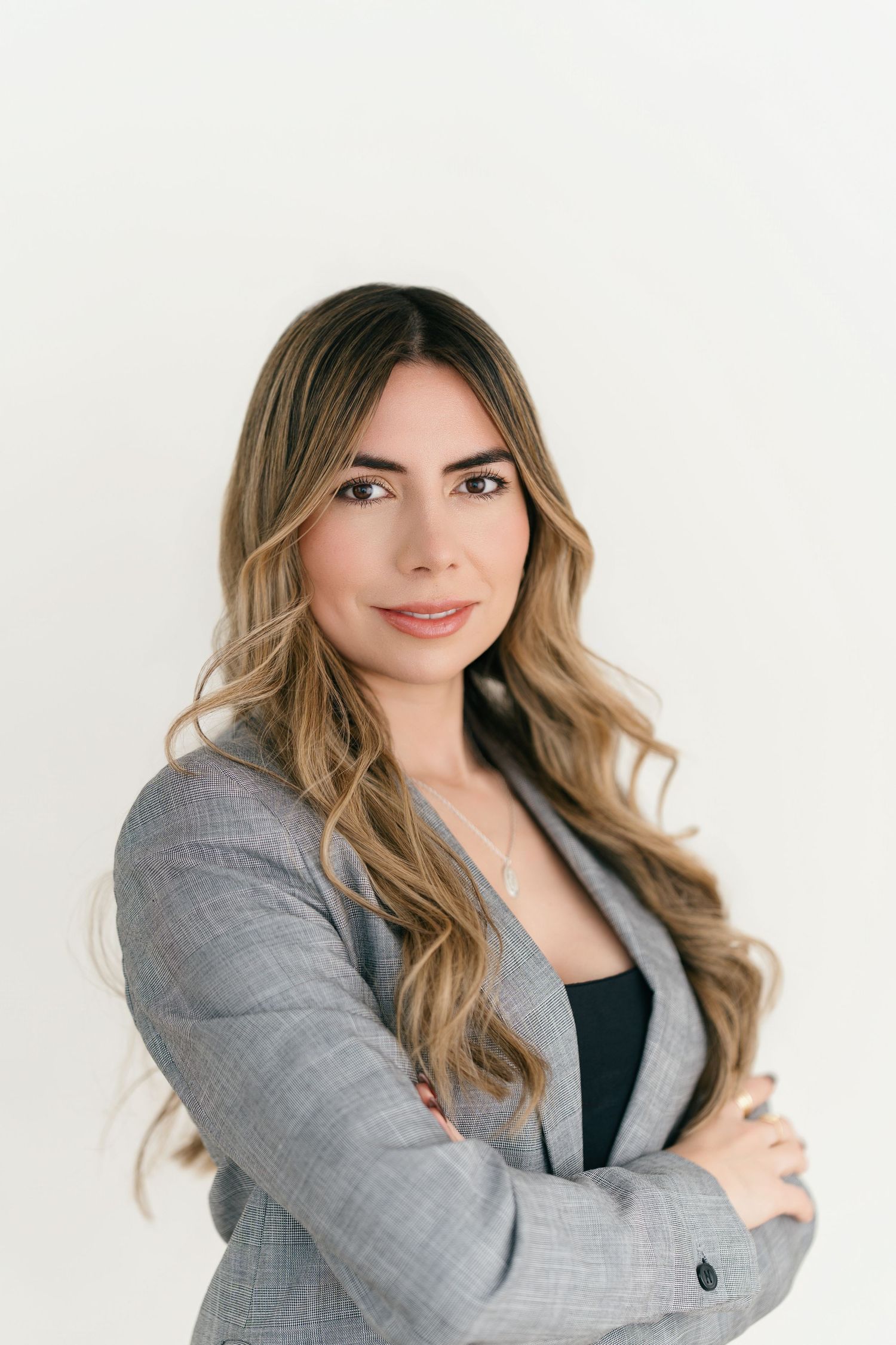 Professional woman in gray blazer with long wavy hair, arms crossed, posing confidently against white background.