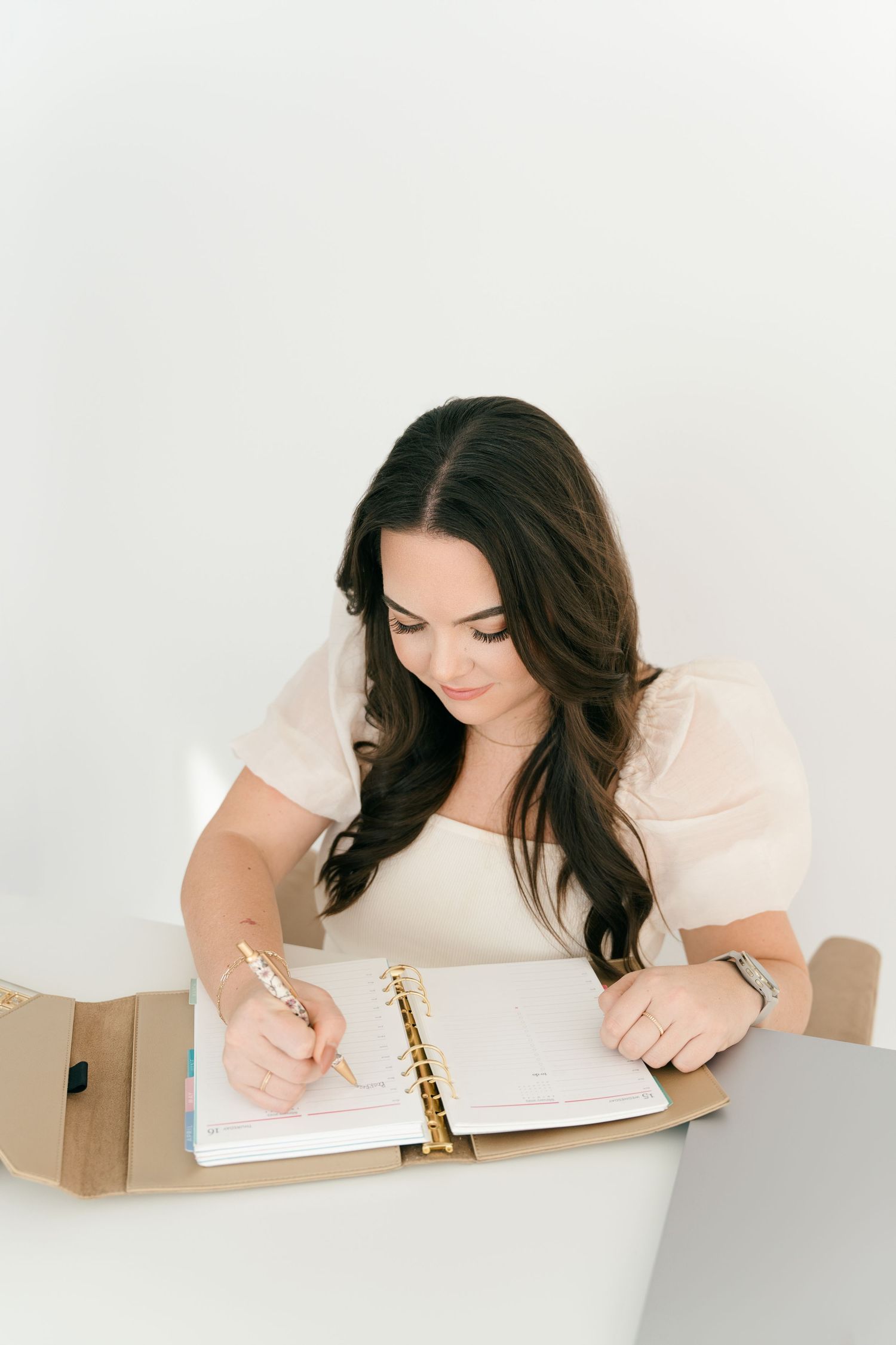 Dark-haired woman in cream top writing in spiral planner with gold pen at white desk.