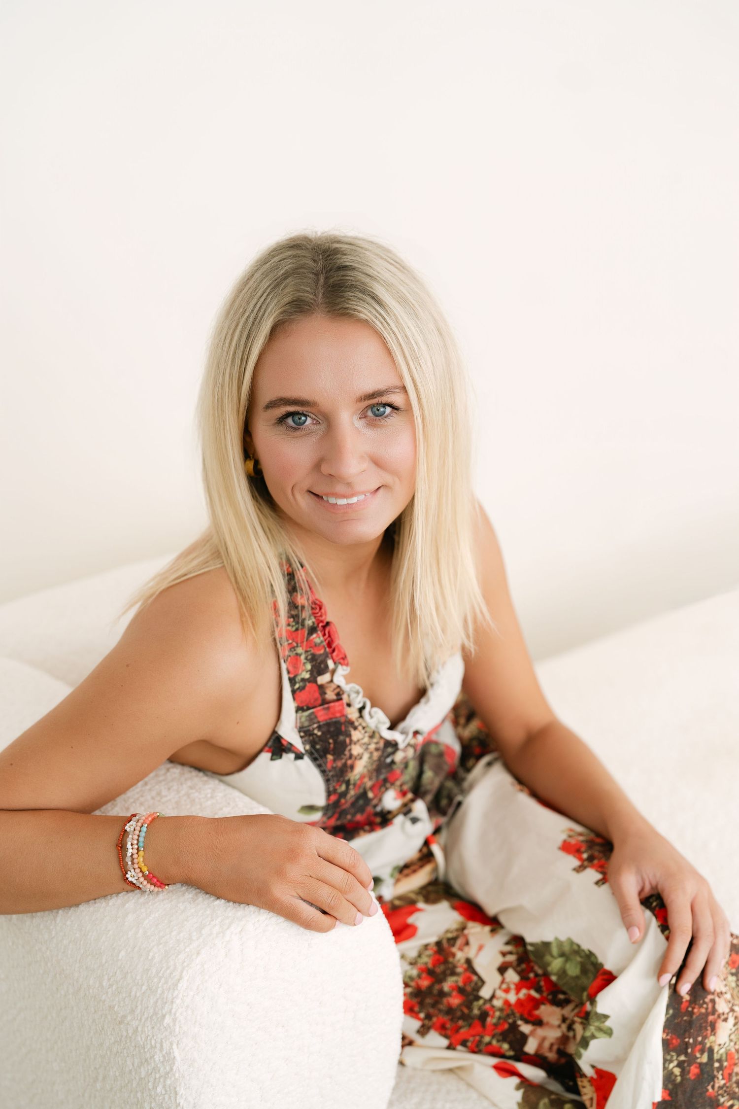 Smiling blonde woman in floral dress sitting on white bed, looking at camera with blue eyes.